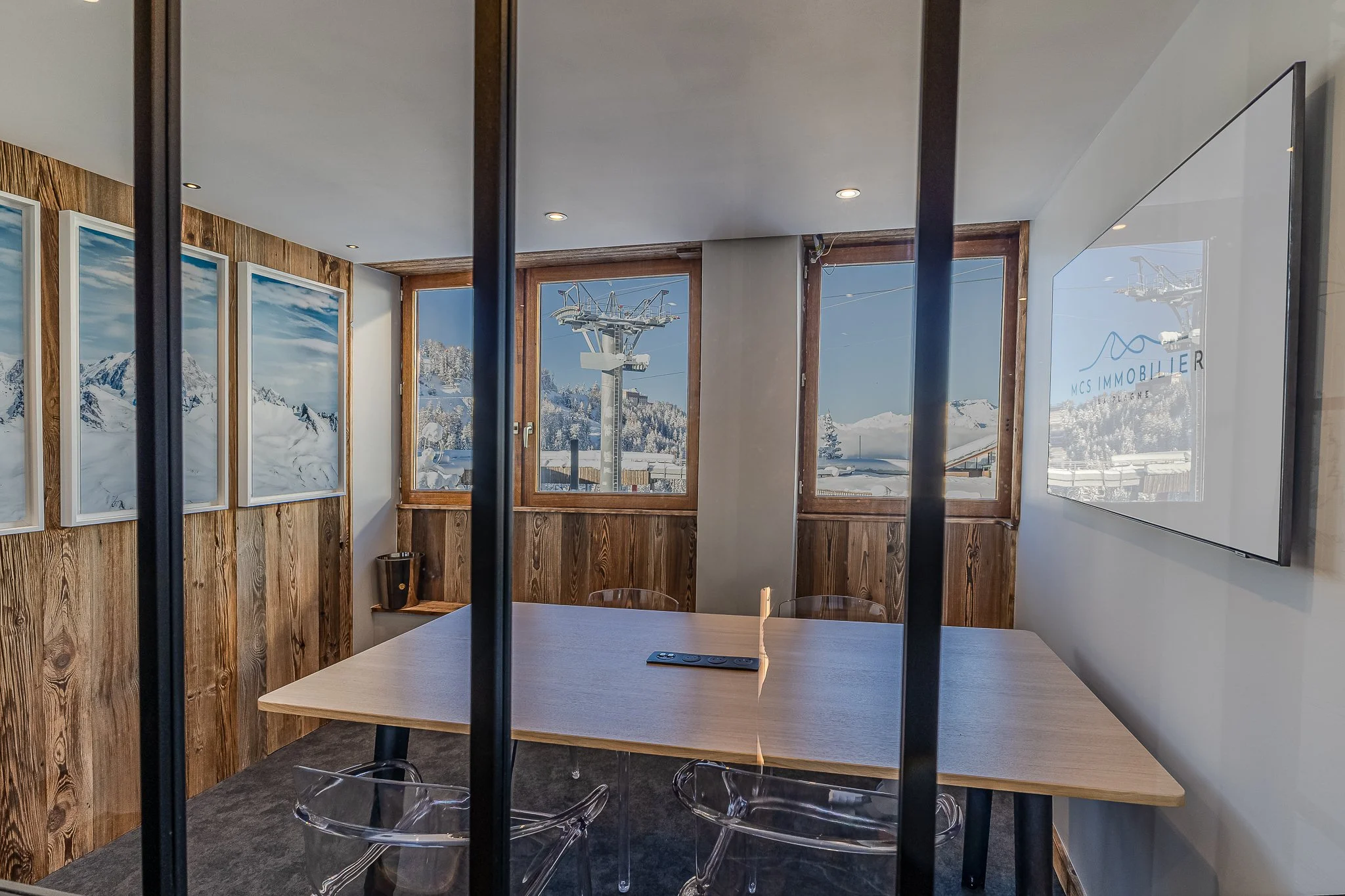 Salle de réunion avec un tableau en bois, plusieurs chaises en plastique transparent, et des fenêtres montrant des paysages enneigés et une station de ski avec des remontées mécaniques.