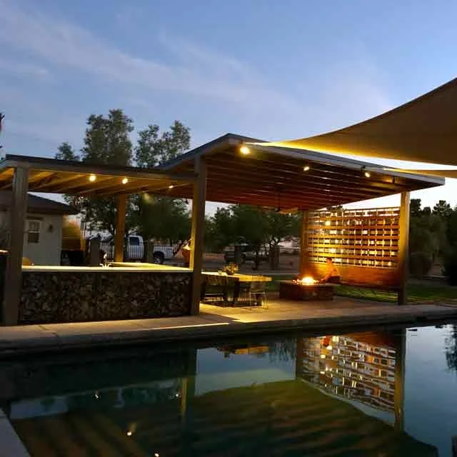 A softly lit patio next to a pool. The patio features a paved floor, custom wooden walls, and an overhead roof.