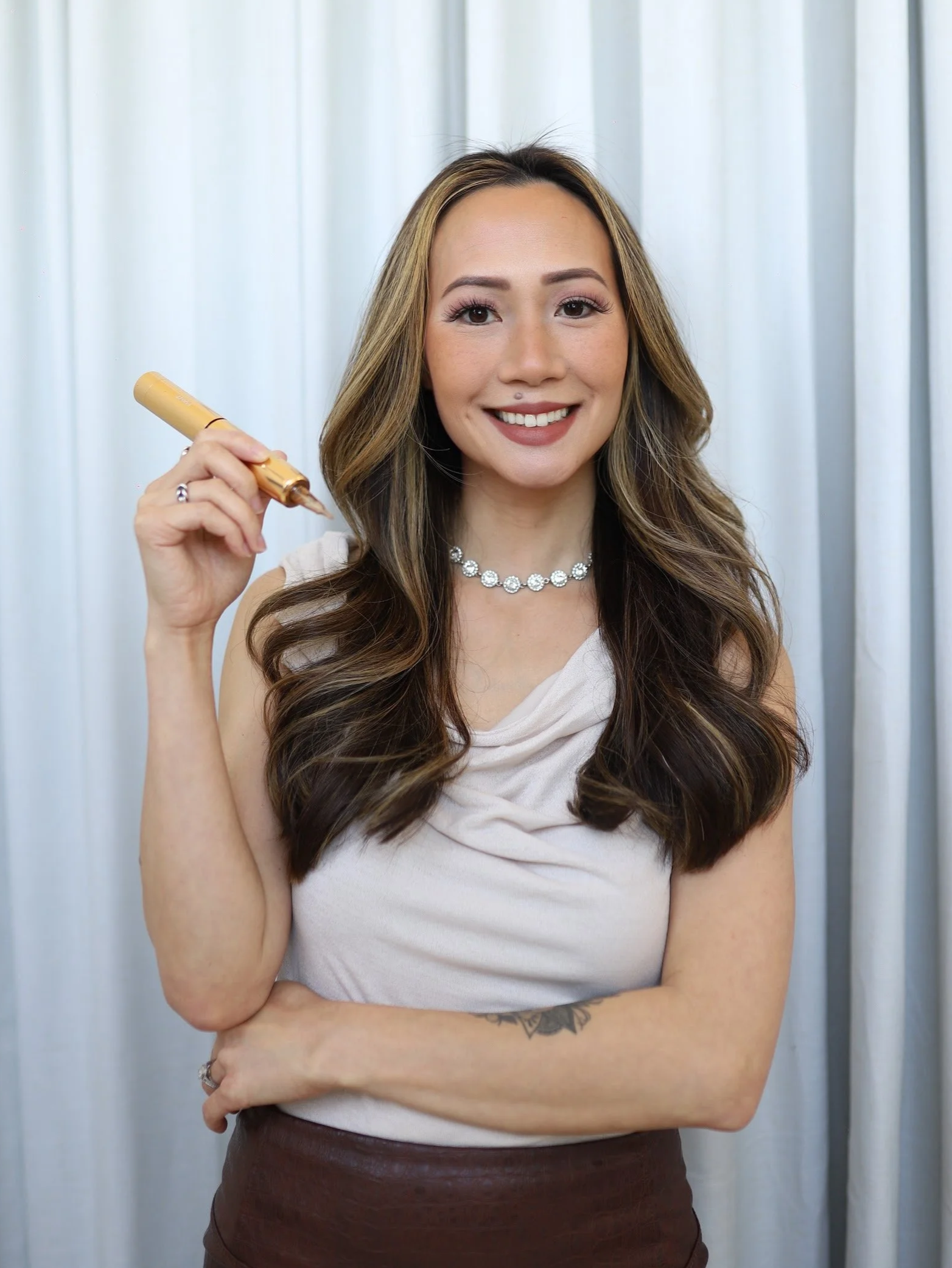 Milan, owner of Brows By Milan, holding a gold-colored tattoo pen in her right hand against a light-colored curtain background.