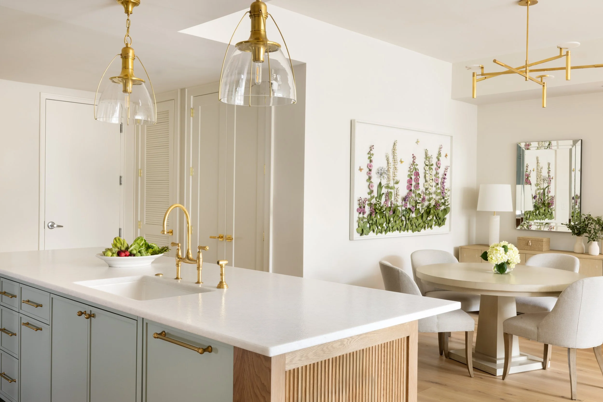Bright and modern kitchen and dining area with white walls, a round dining table with white chairs, a floral wall art, a mirror, a white table lamp, and a bunch of flowers on the table. Gold accents include pendant lights, a faucet, and cabinet handl