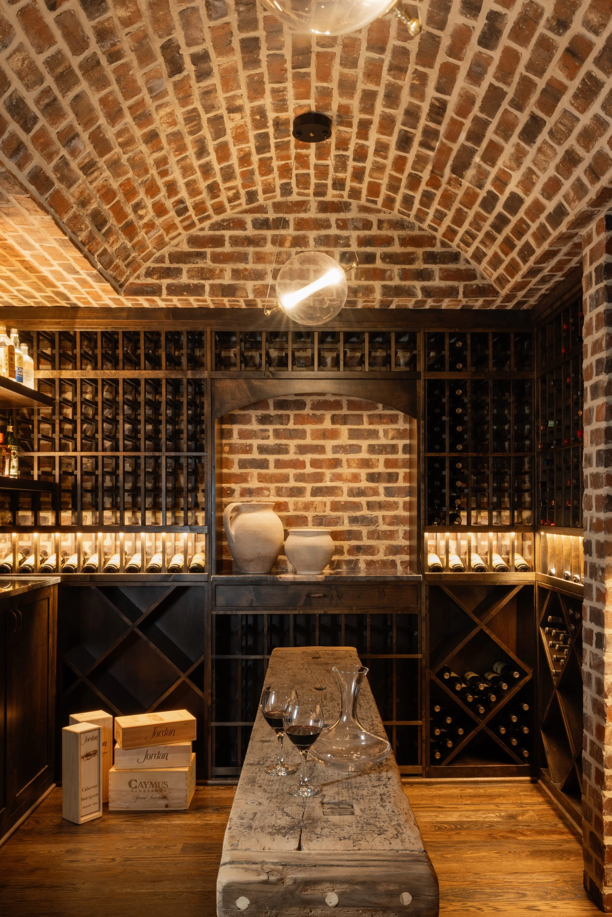 An elegant wine cellar with brick arch ceiling, wooden wine racks filled with bottles, a rustic wooden table with glasses of red wine and a glass decanter, decorative pottery on shelves, and wood crates on the floor.