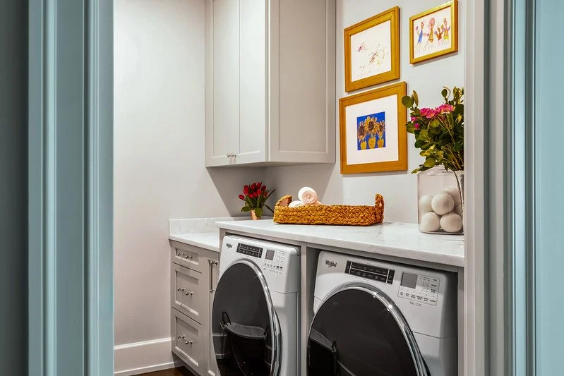 Laundry room with white cabinetry, a countertop, two front-loading washing machines, and wall art with colorful frames.