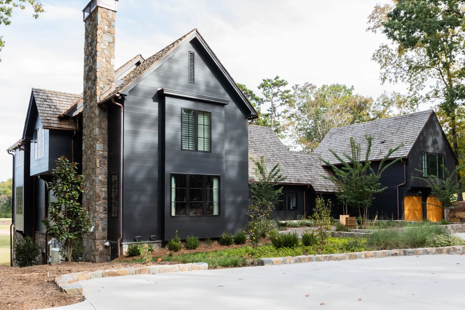 Modern custom home built by Will Johnson Building Company featuring a black brick exterior, steep gabled rooflines, tall chimney detail, large black-framed windows, and natural landscaping integrated into a quiet residential setting.