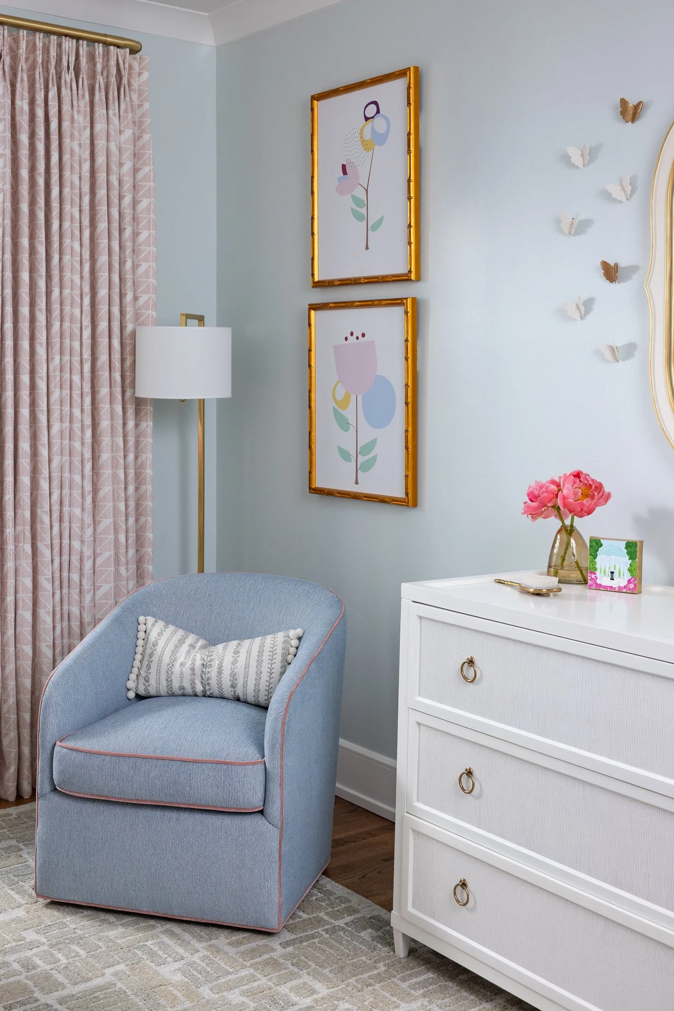 A cozy corner of a bedroom with a light blue armchair, a white dresser, pink peony flowers in a glass vase, and wall art with geometric floral designs. Light pink curtains with a subtle pattern are on the window, and small butterfly decorations are o