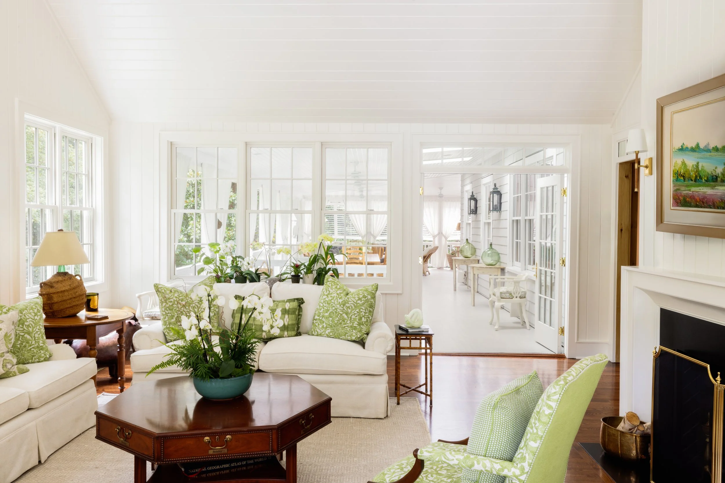 Bright living room with white walls, large windows, green and white cushioned furniture, a wooden coffee table with a potted plant, and a fireplace.