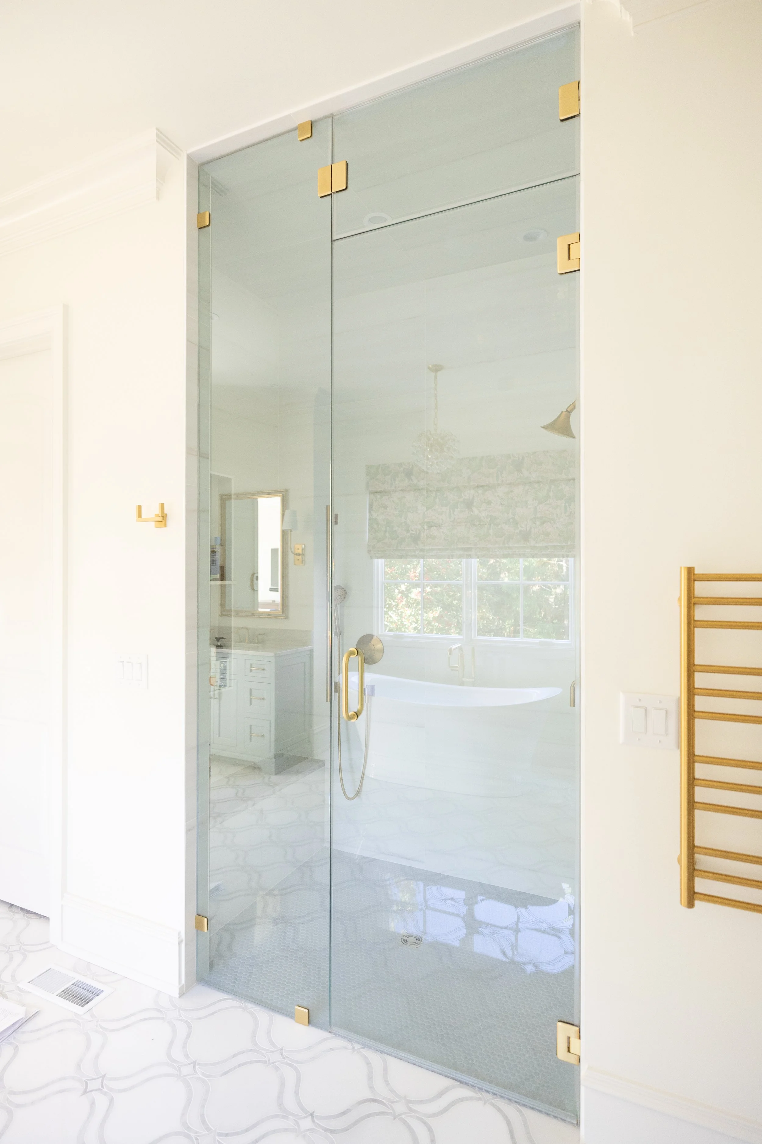 A bathroom with a glass shower enclosure, gold hardware, a tub visible through the glass, a window with a floral shade, a chandelier, and bathroom fixtures reflected in the glass.