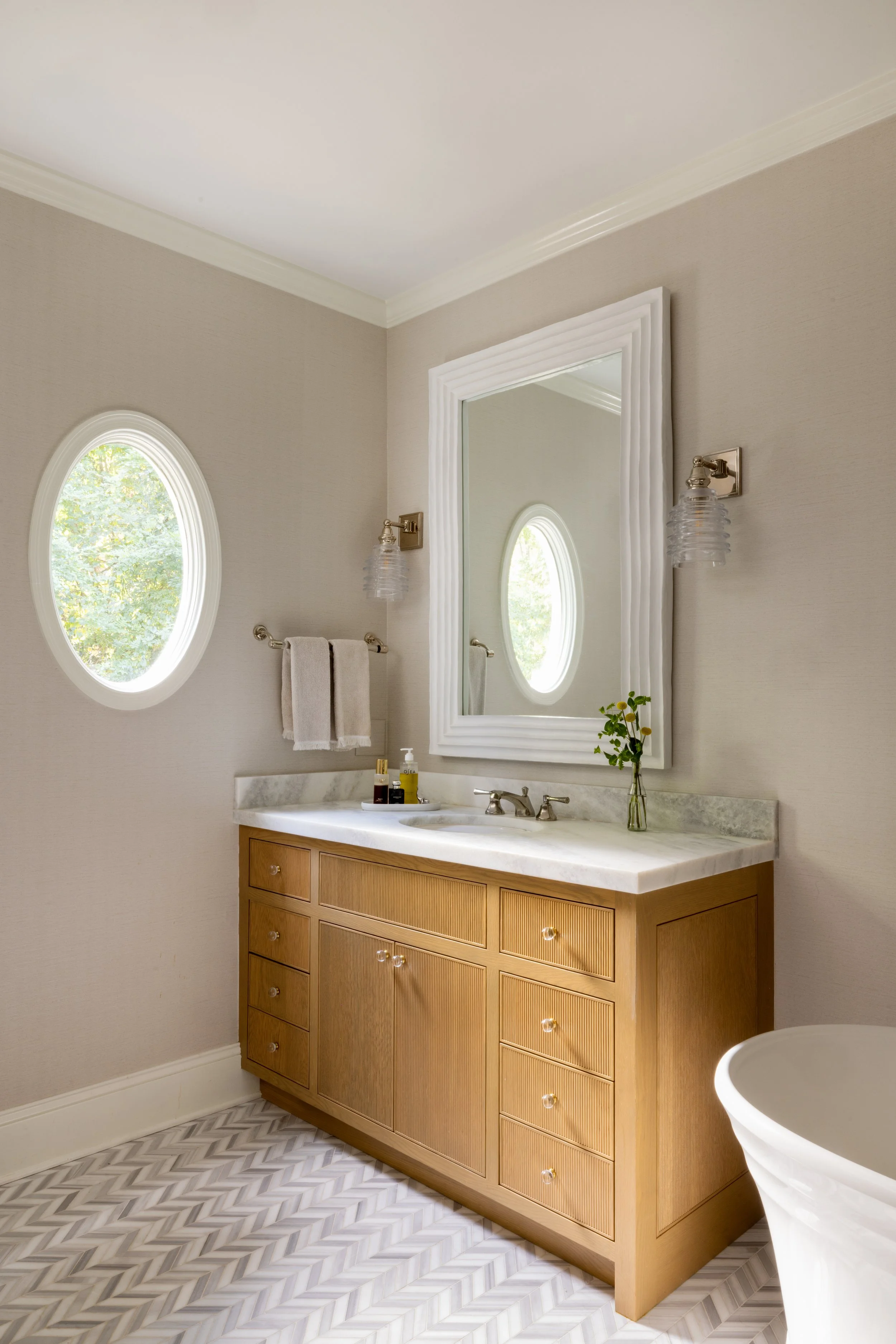 Bathroom with a wooden vanity, marble countertop, large mirror, wall-mounted light fixtures, oval window, and a vase with yellow flowers.