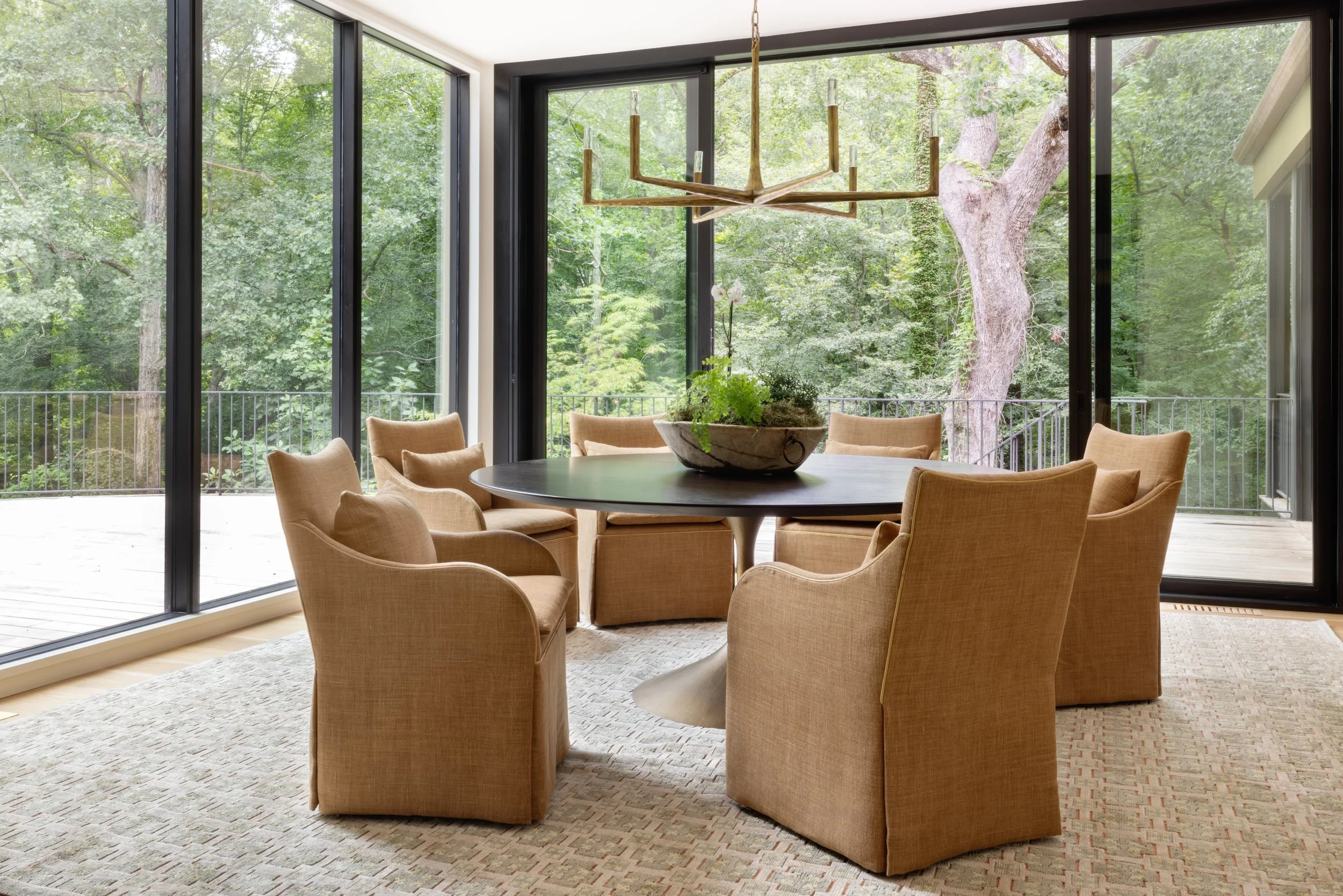 A dining room with a round table surrounded by eight beige upholstered chairs. There is a large bowl with plants on the table. The room has floor-to-ceiling glass sliding doors showing a wooden deck and green trees outside. A modern chandelier hangs 