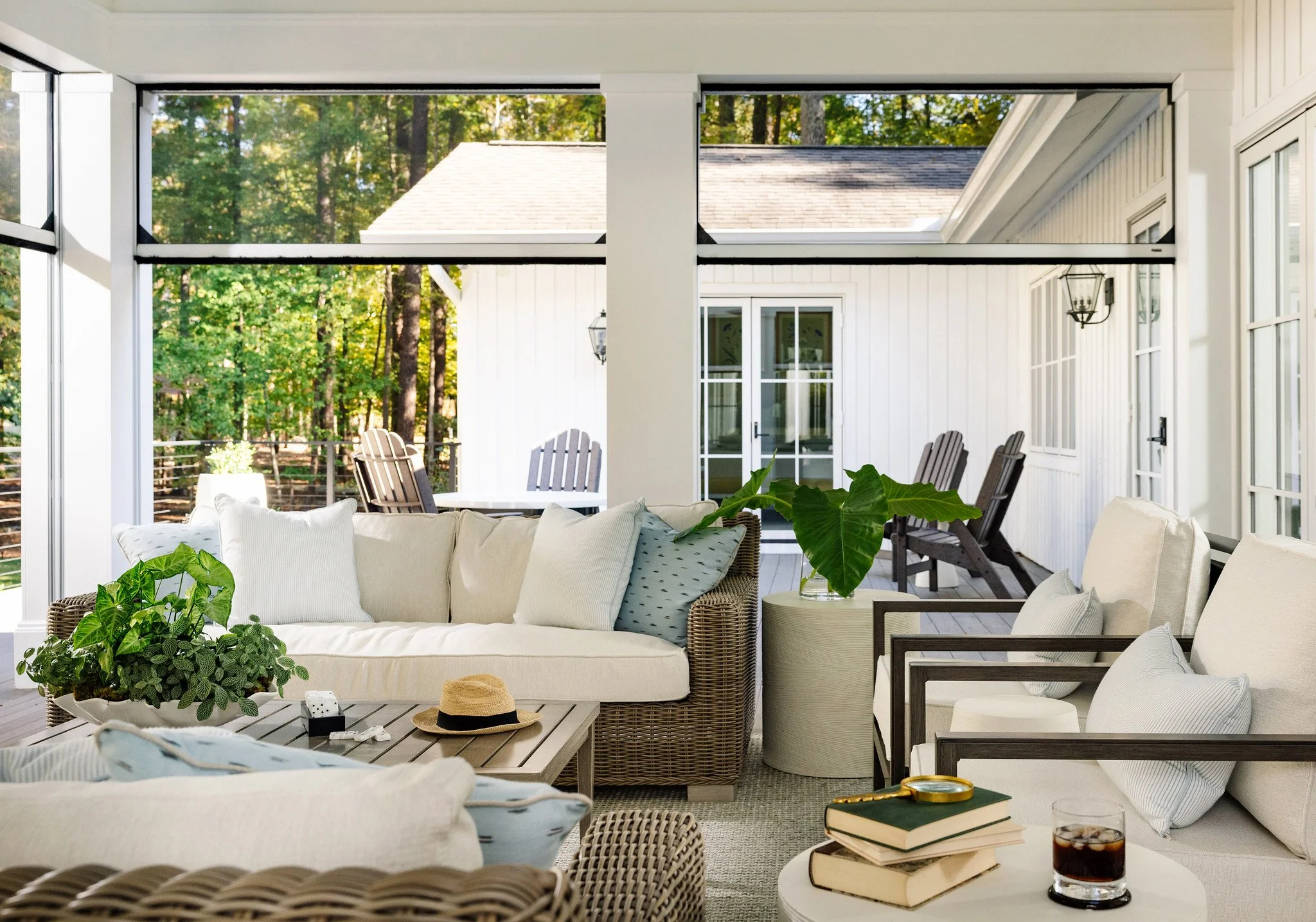 Sunroom with outdoor view, beige couches and armchairs with pillows, potted plants on tables, hat on coffee table, books, glass of soda, and large windows showing a deck with chairs and a background of trees and a house.