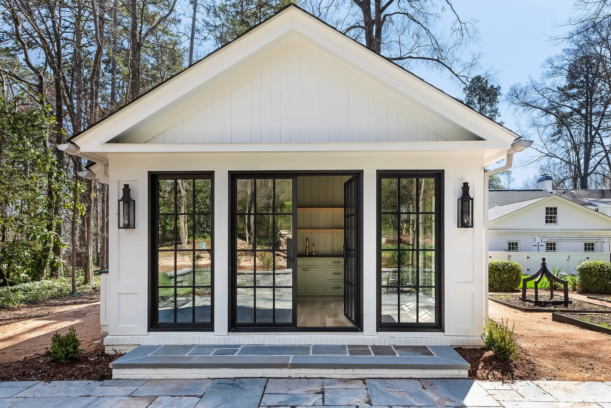 A white house with black-framed glass doors leading to a small patio, surrounded by trees and shrubbery.