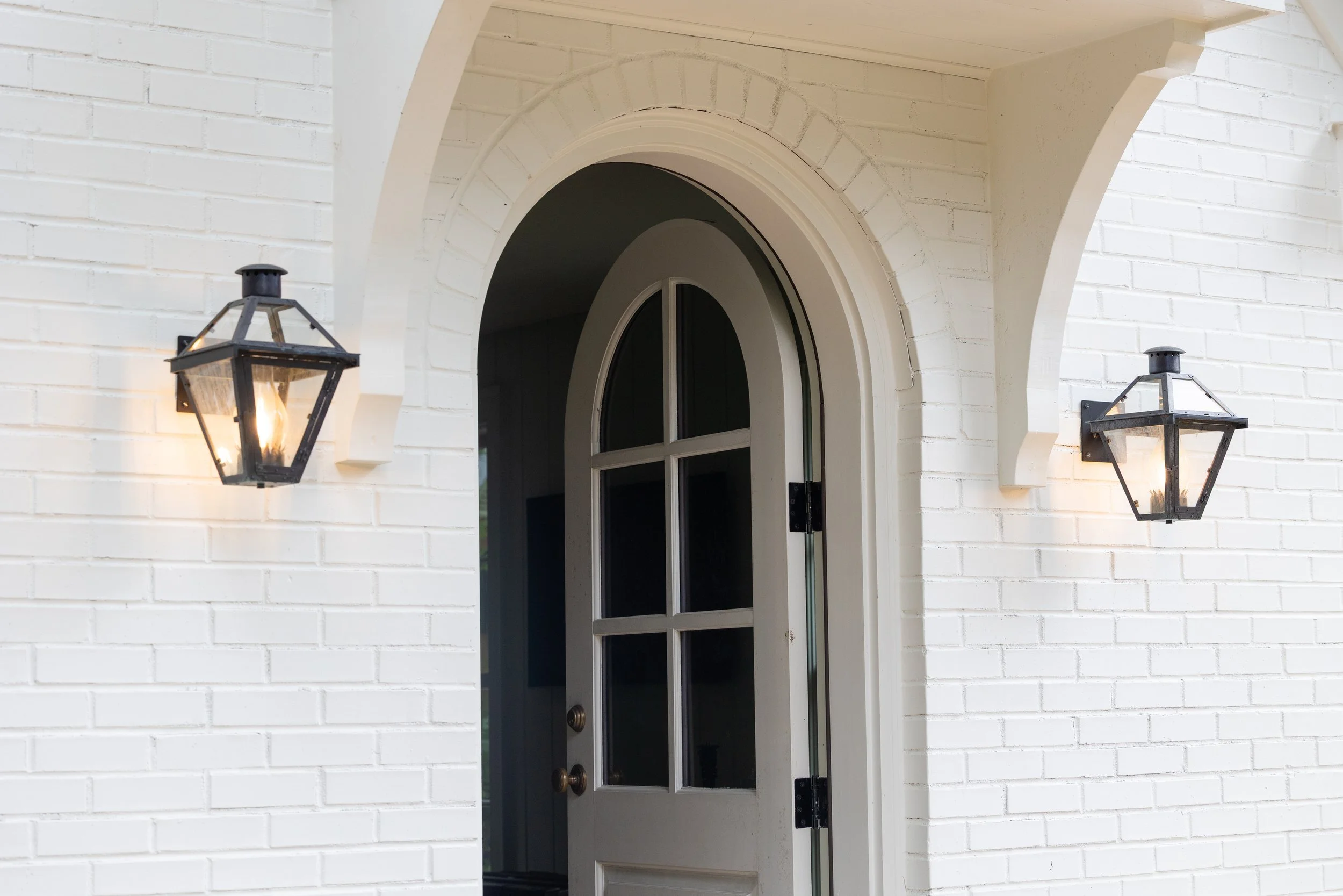 White brick house with an arched front door, flanked by two black lantern-style wall lights.