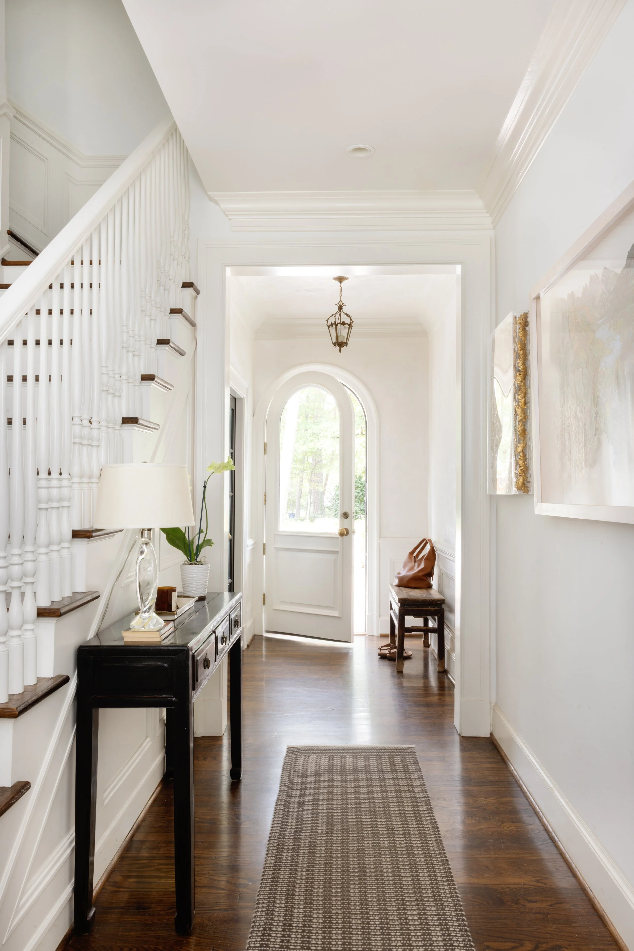 Bright hallway with white walls and wood flooring, featuring a small dark console table with a lamp and potted plant, an abstract painting on the wall, a bench with a bag, and an arched window at the front door.
