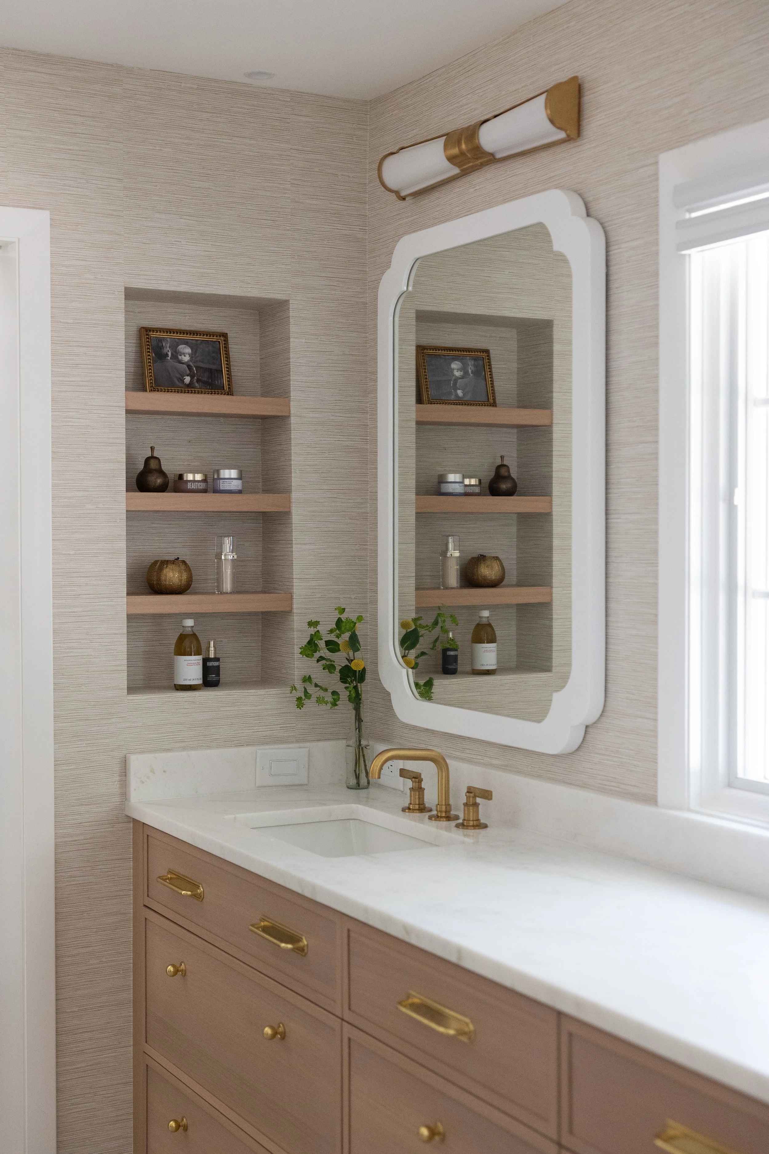 A bathroom vanity with pink drawers, a white marble countertop, and gold hardware, reflected in a white framed mirror, with a vase of greenery and shelves with decorative items.