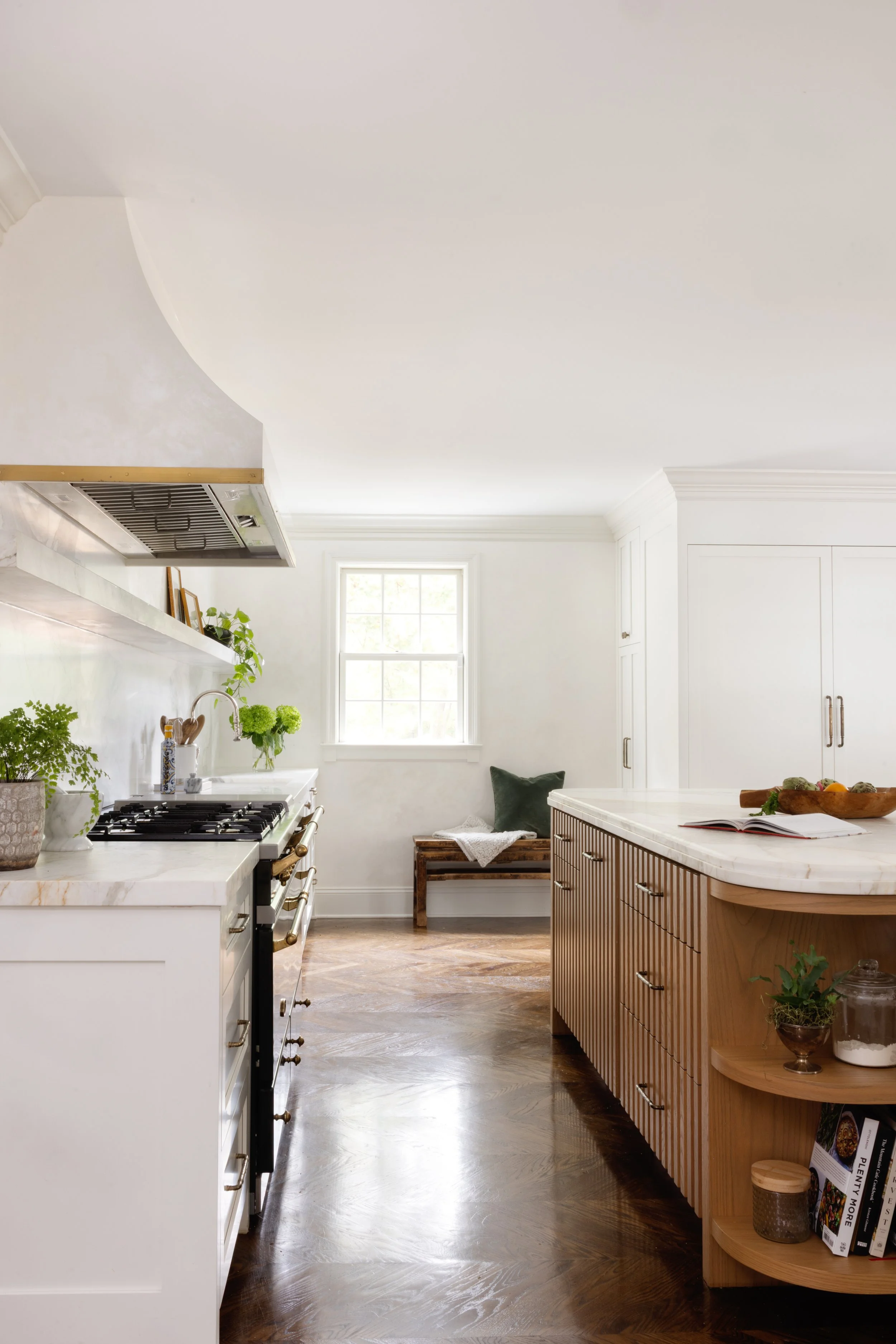 Bright kitchen with white walls, natural wood island, white cabinetry, dark hardwood floors, plants, and a window with a seating area.