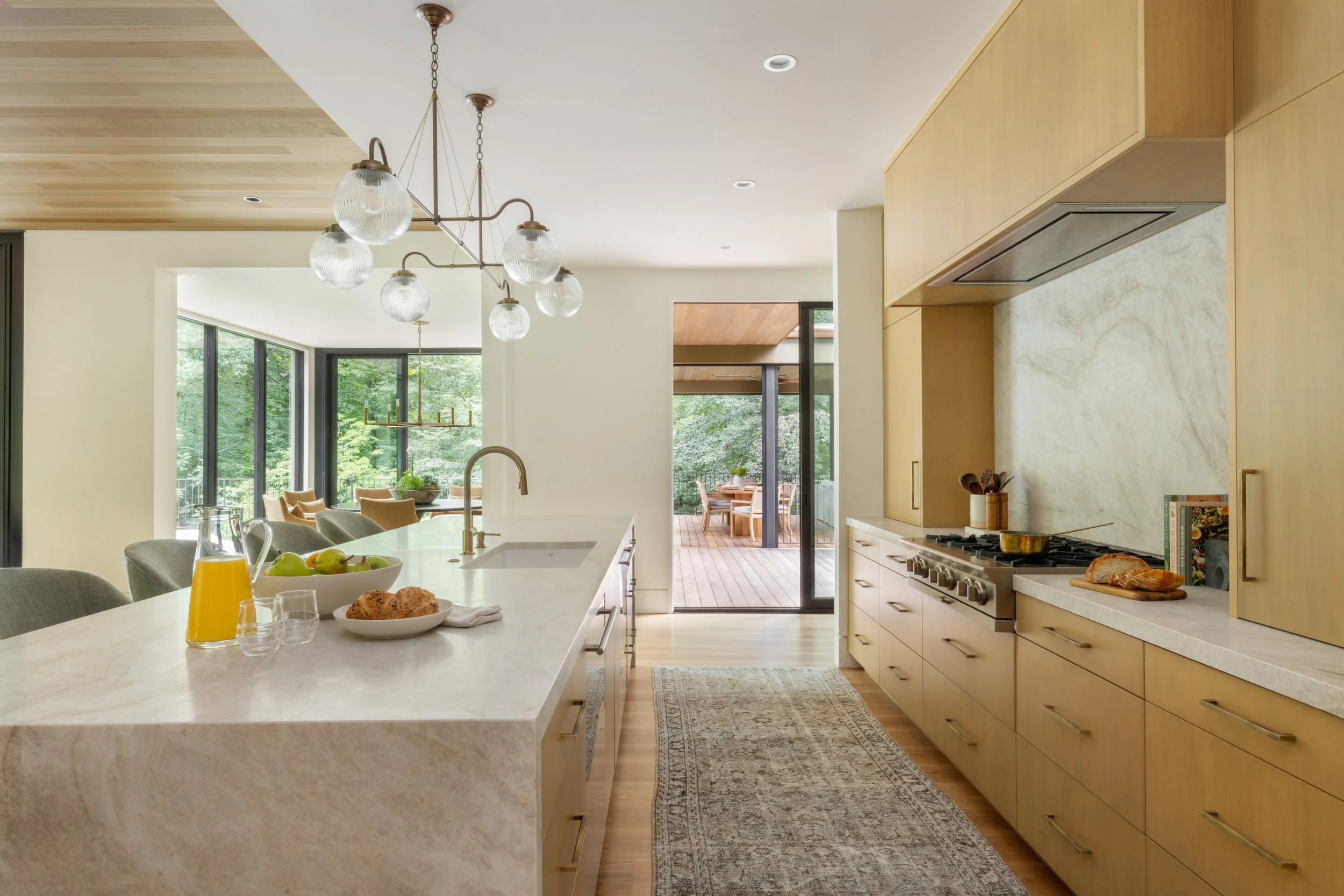 Modern kitchen with light wooden cabinets, marble countertops, an island with breakfast and snacks, and a view of a deck with outdoor dining, greenery visible through large windows.