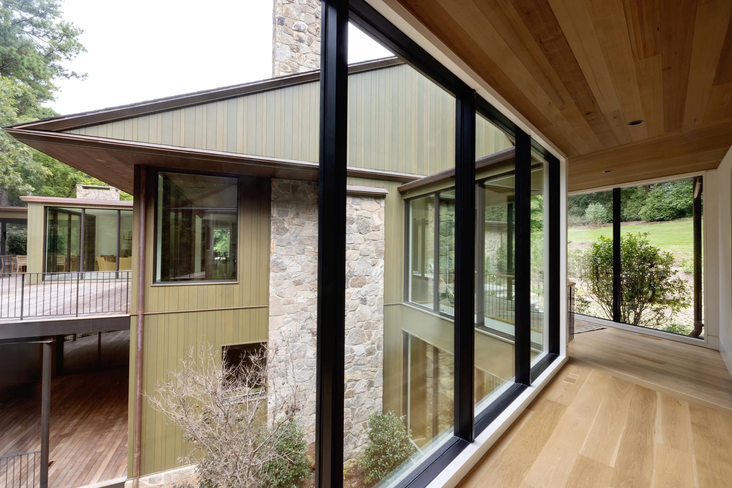 Interior view of a house with large glass windows and wooden ceiling, overlooking an outdoor deck and green yard.