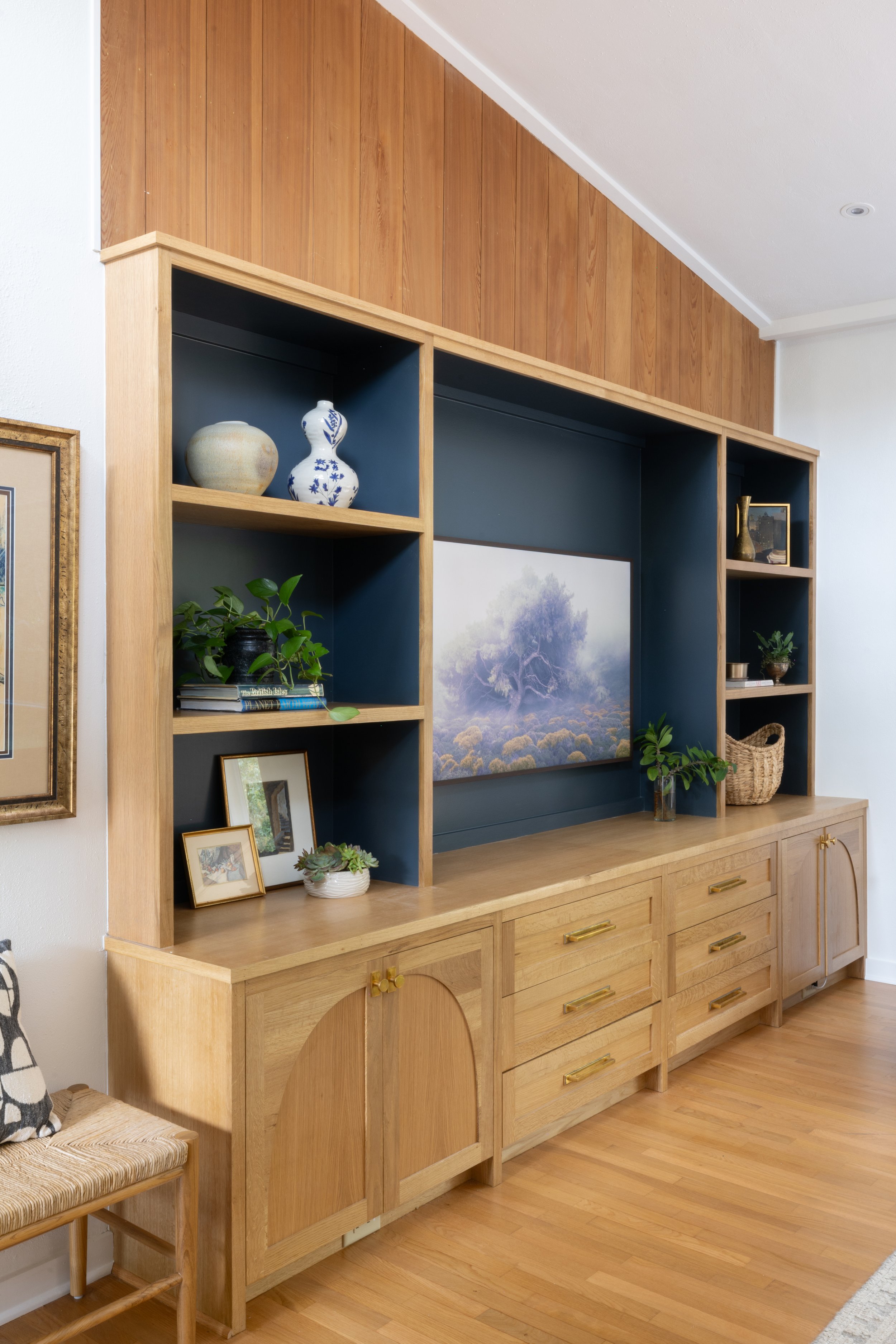 Wooden entertainment center with shelves and cabinets, decorated with vases, framed photos, and plants, with a large landscape painting in the center.