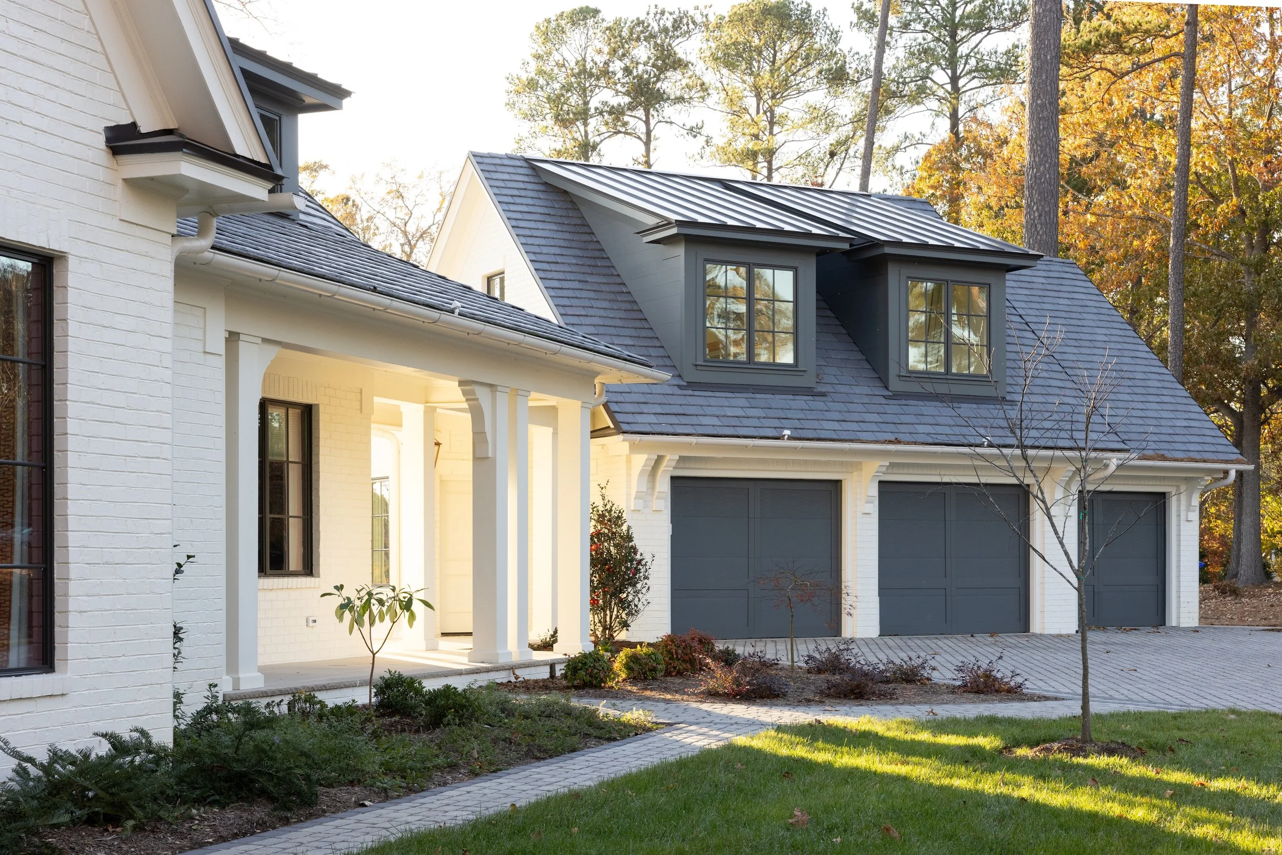 A modern house with white brick exterior, gray garage doors, and dark gray dormer windows, surrounded by a landscaped yard with trees and a paved driveway.