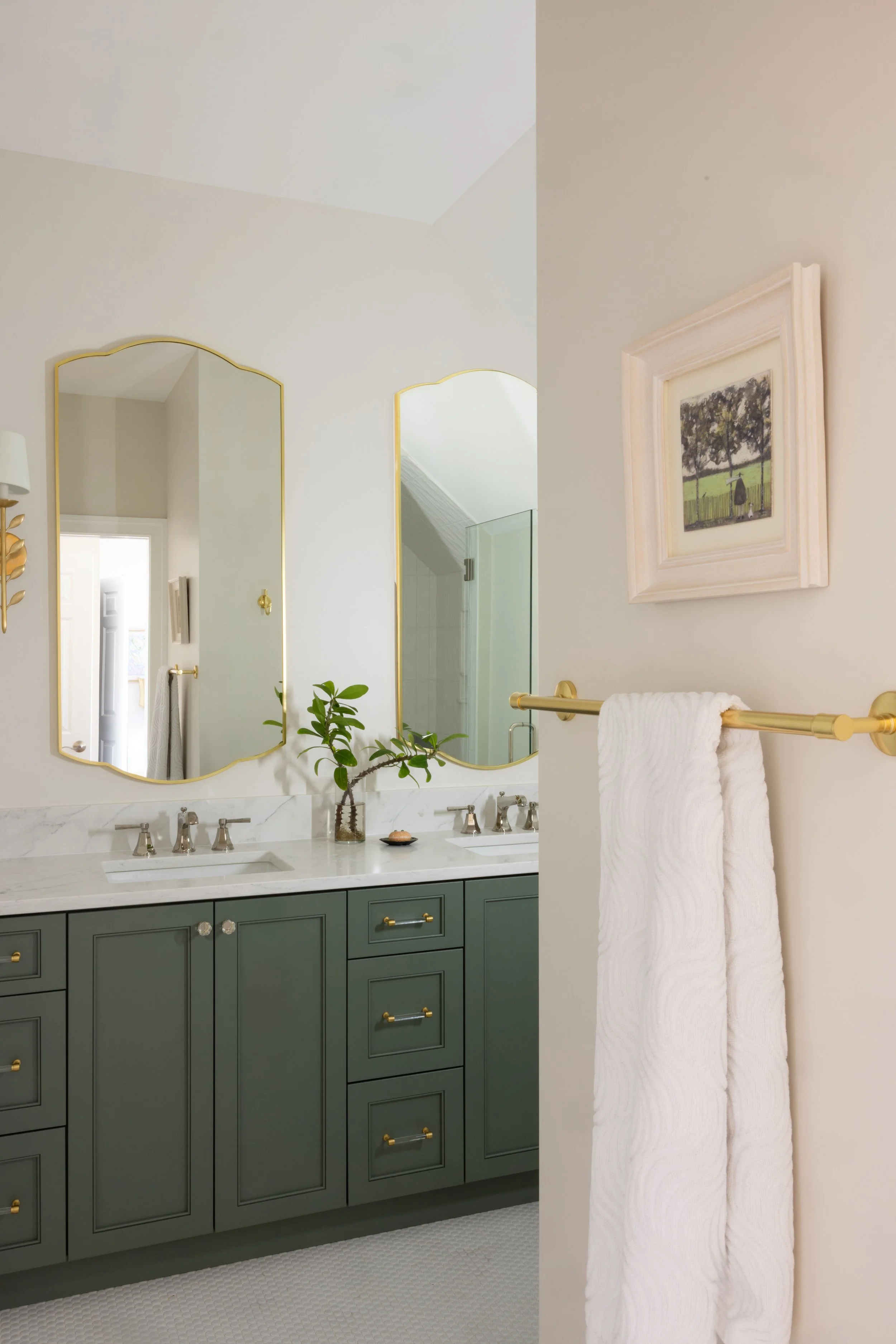 A bathroom vanity with green cabinets, marble countertop, and two sinks with silver faucets, decorated with a small potted plant, framed artwork, a gold towel rack with a white towel, and two gold-framed mirrors above the sinks.