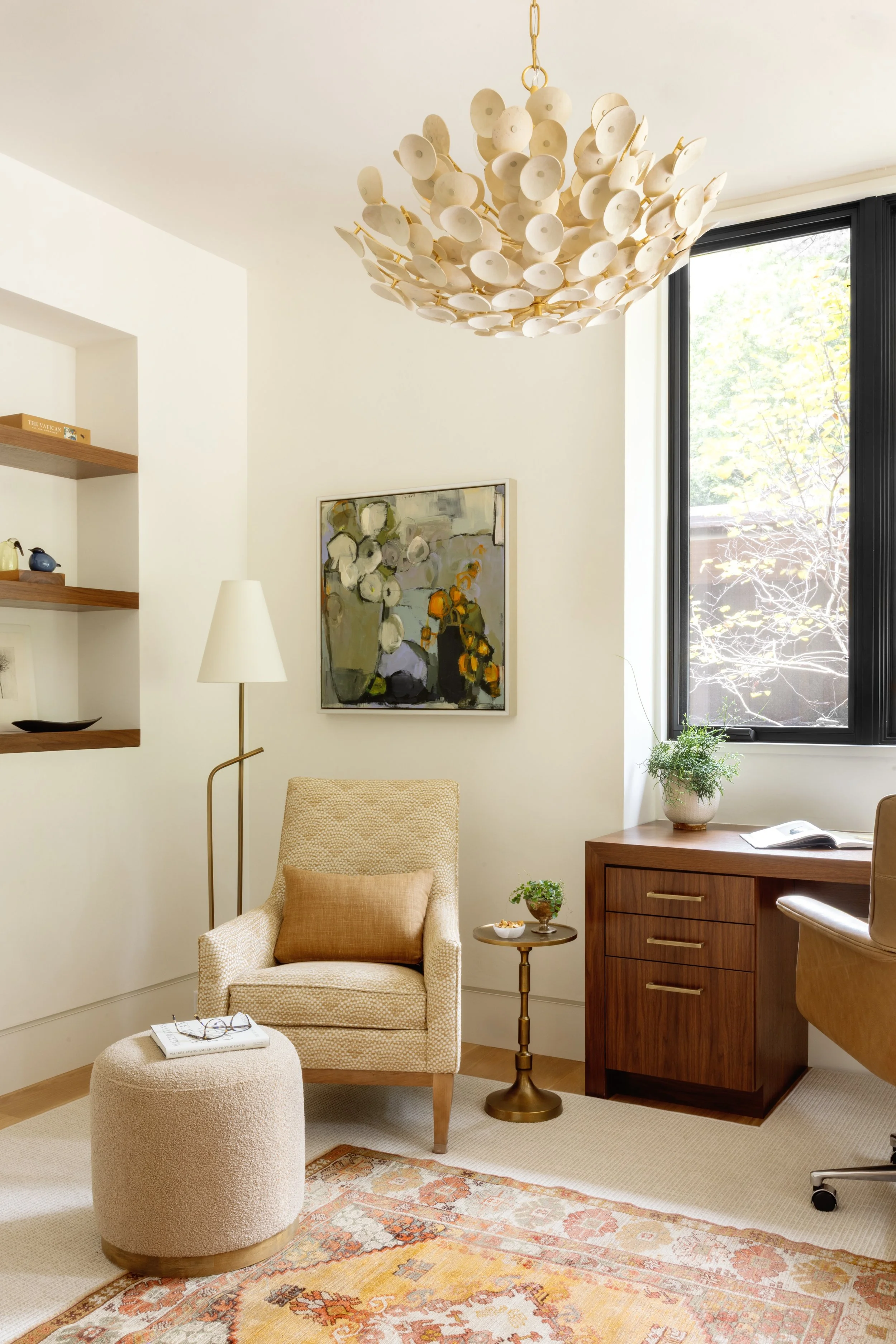 A cozy living space with a beige armchair and ottoman, a small side table with a plant, a wooden desk with a plant and book, a patterned rug, a large window, and a modern chandelier hanging from the ceiling.