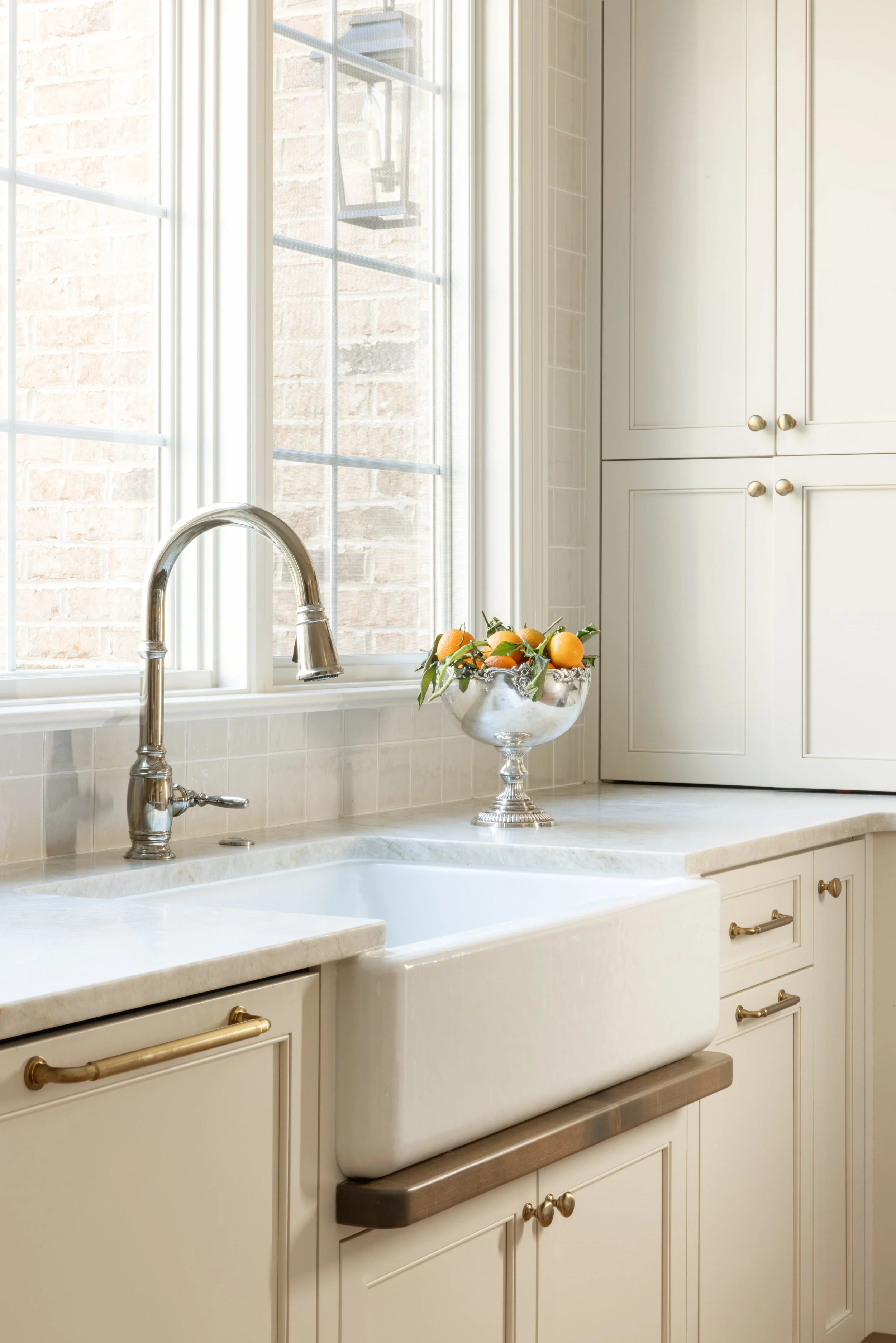 A farmhouse kitchen with a white apron sink, silver faucet, cream cabinets with gold handles, a window with white trim, and a silver fruit bowl containing oranges.