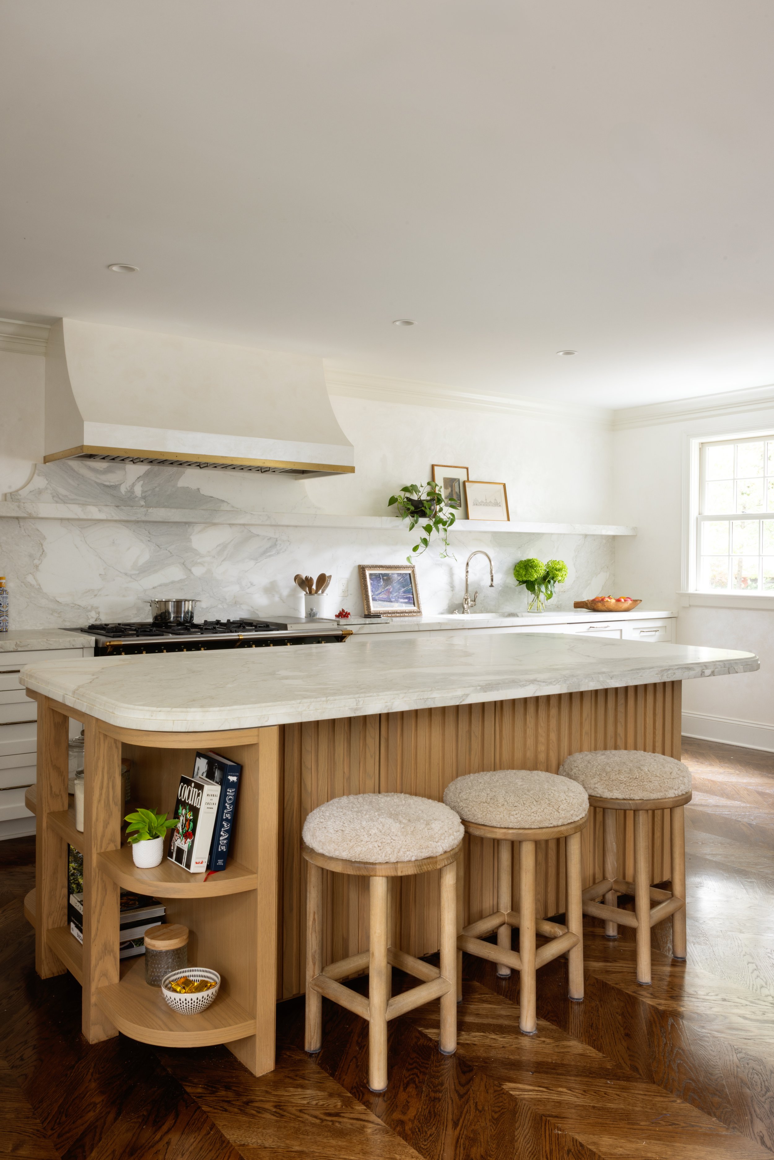 Modern kitchen with white marble countertops, wooden island with shelves, three beige cushioned stools, and a window letting in natural light.