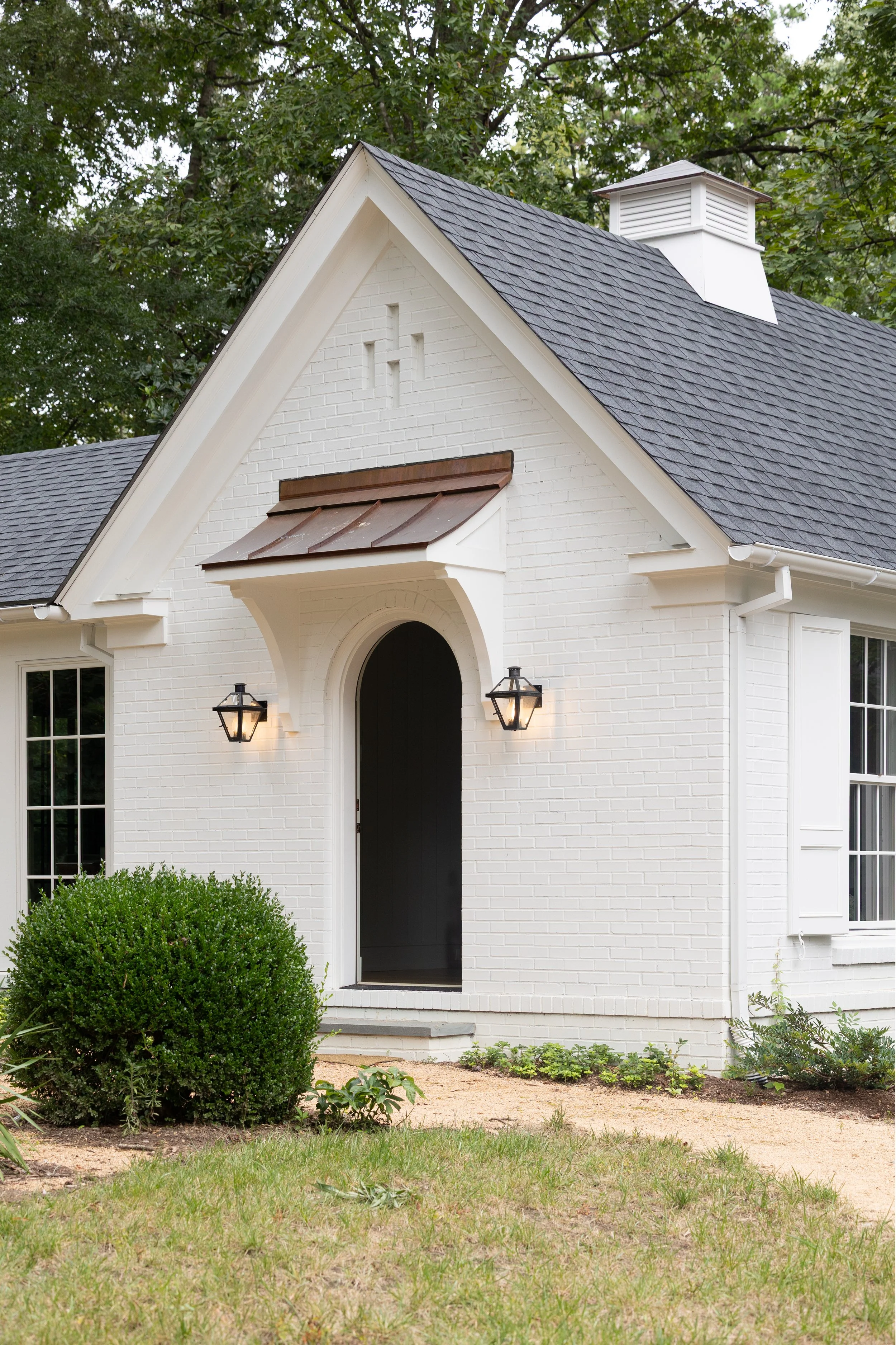 White house with black front door, two wall-mounted lantern lights, a small bush in front, and a grey shingled roof surrounded by green trees.