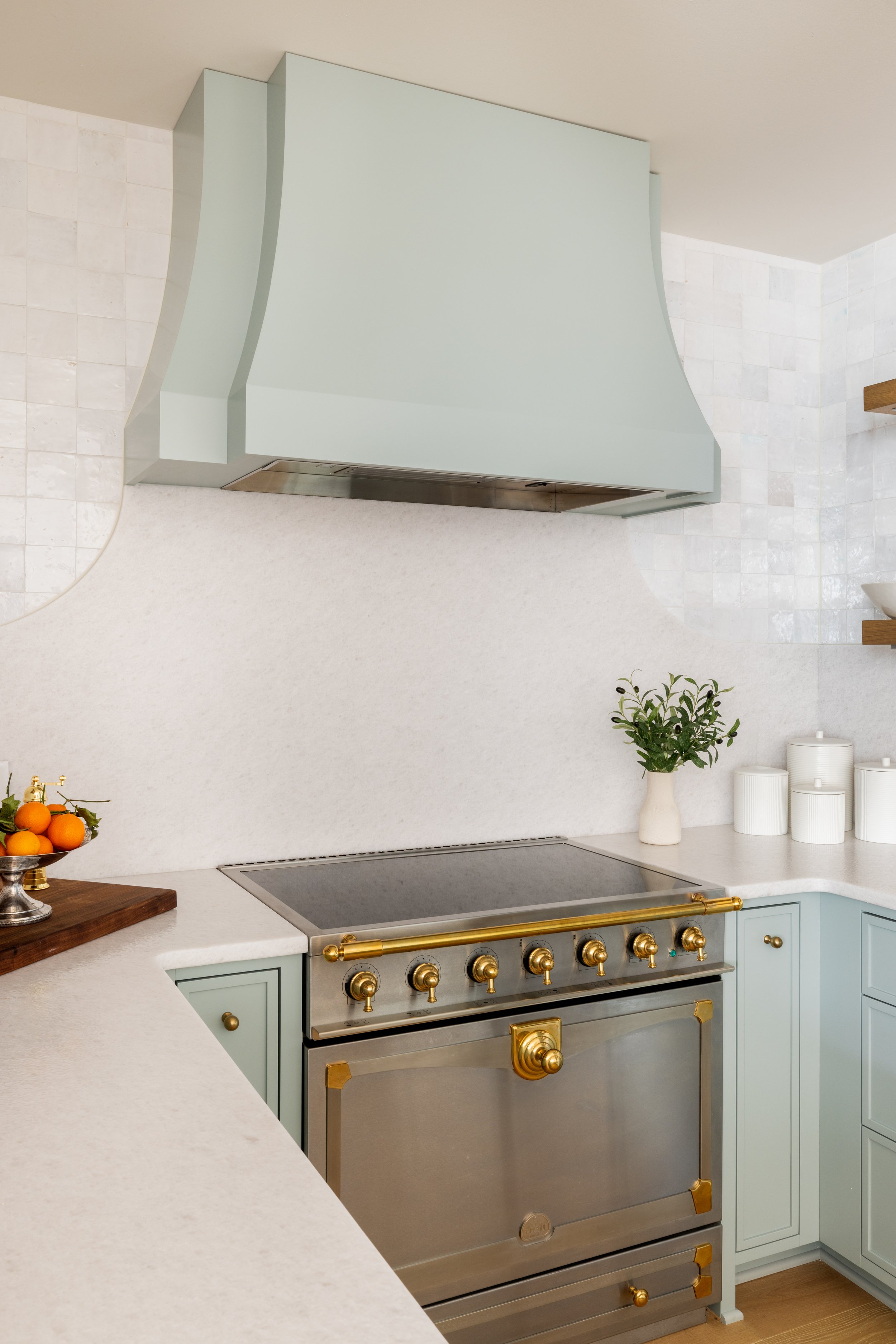 Modern kitchen with pastel blue cabinets, a stainless steel stove with gold accents, white countertops, a minimalist vase with greenery, and a range hood.