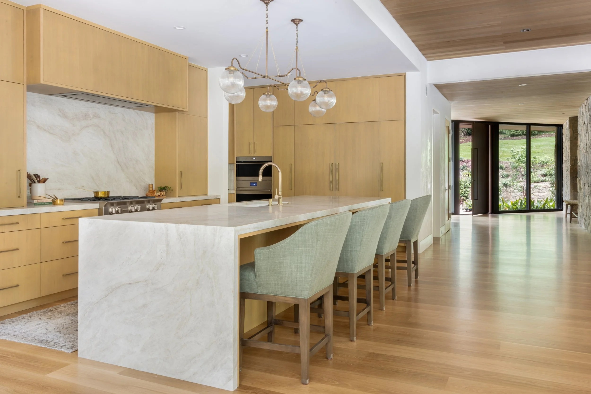 Modern kitchen with light wood cabinets, marble island with a sink, and green upholstered bar stools. Large doors lead to an outdoor garden area.