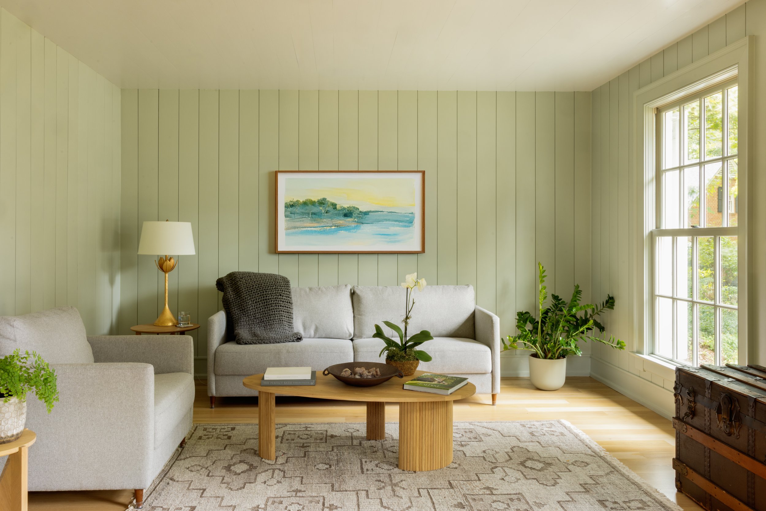 Living room with light green paneled walls, large window, cream sofa with gray throw pillow, beige armchair, wooden coffee table with books and orchid, floor lamp, potted plant, framed landscape painting, patterned rug, and vintage chest.