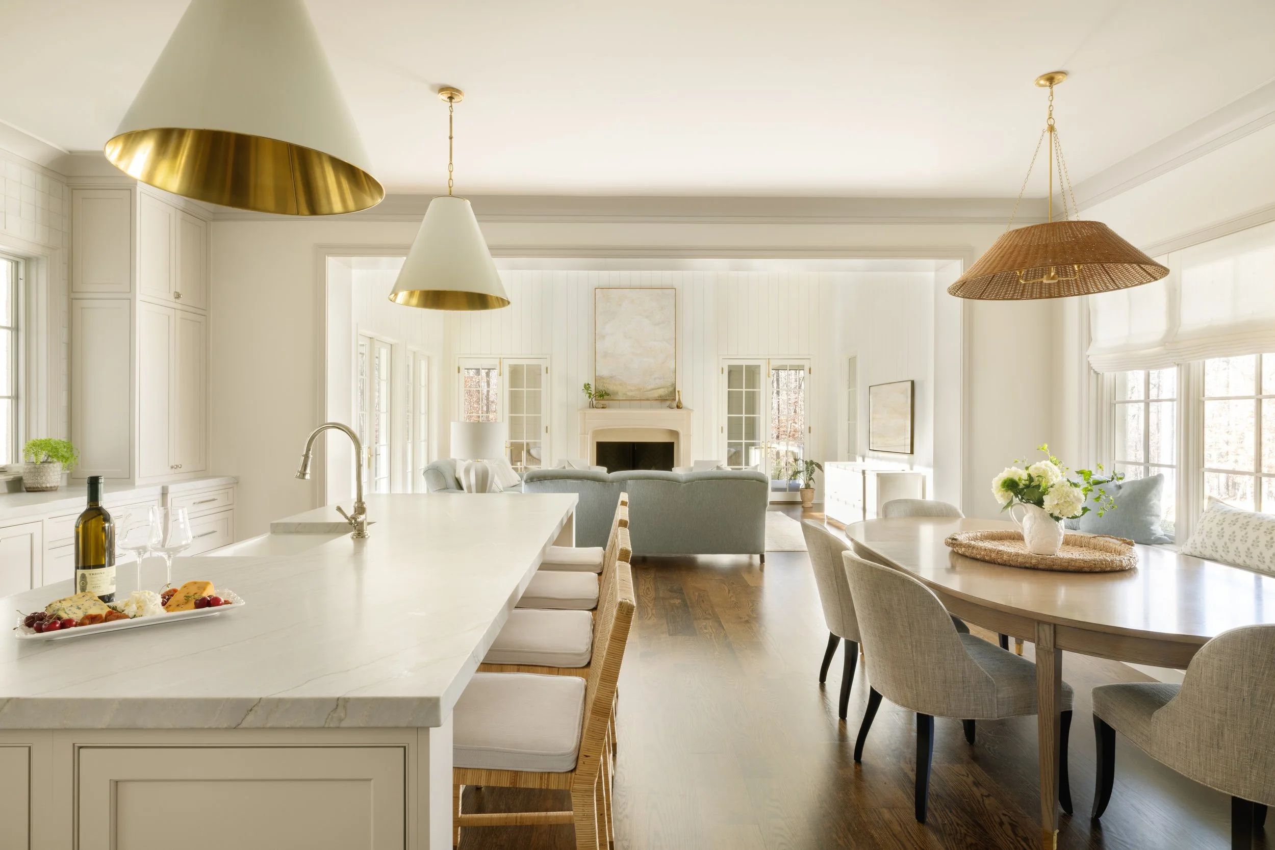 Bright, open-concept kitchen and living room with white cabinetry, a large marble kitchen island, and a dining area. The living area has a fireplace, a sofa, and large windows letting in natural light.