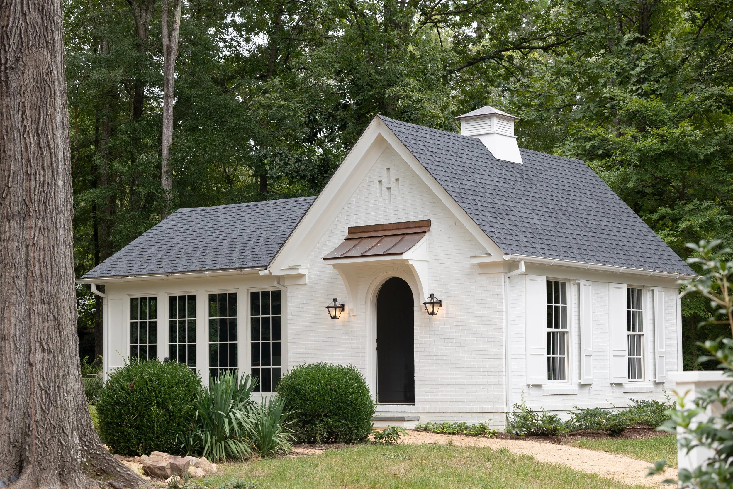 A white house with a gabled roof, black shutters, and a small front porch, surrounded by greenery and trees.