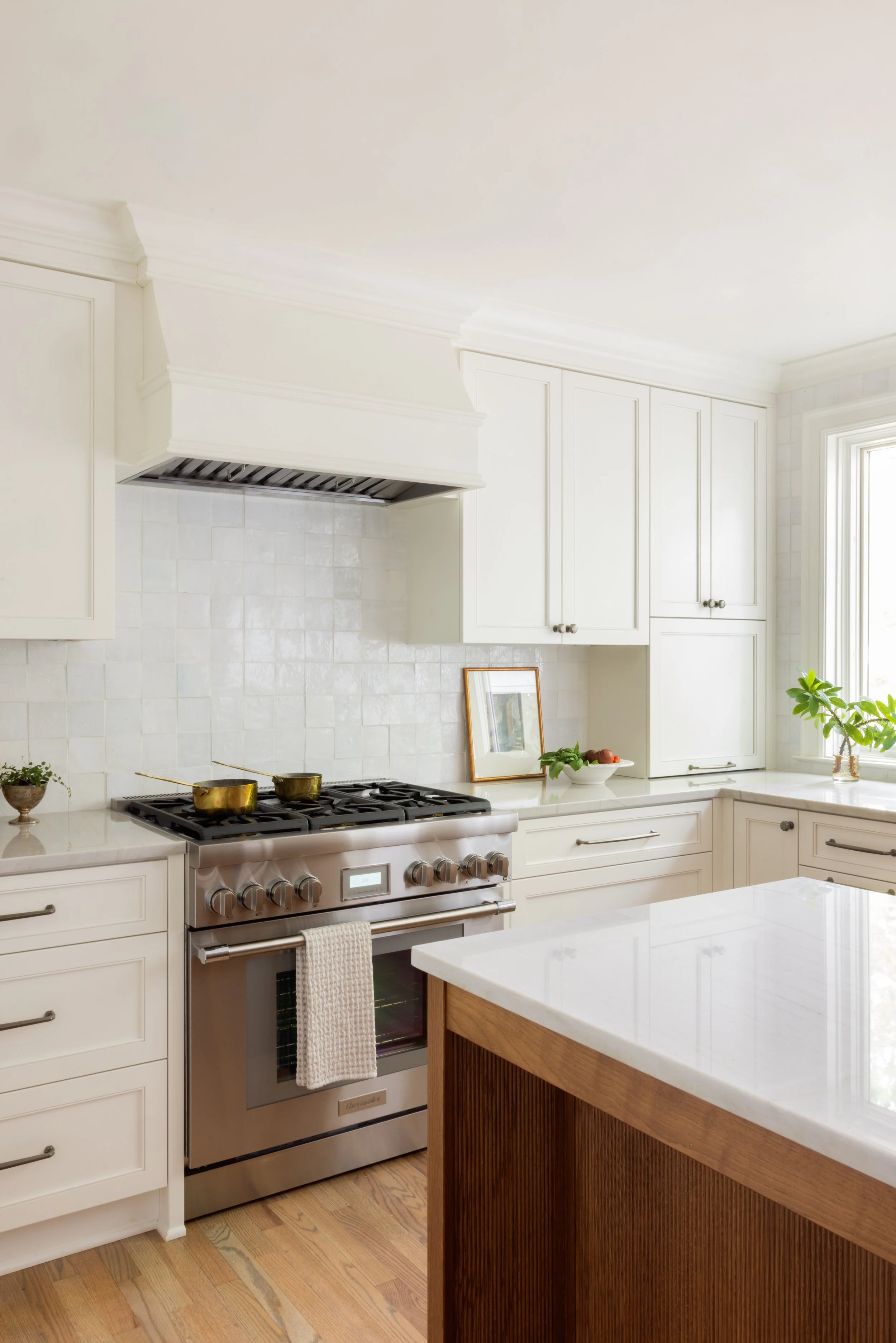 A modern kitchen with white cabinets, a stainless steel oven and stovetop, and a white marble countertop island. Potted plants and framed artwork are on the counters near a window.