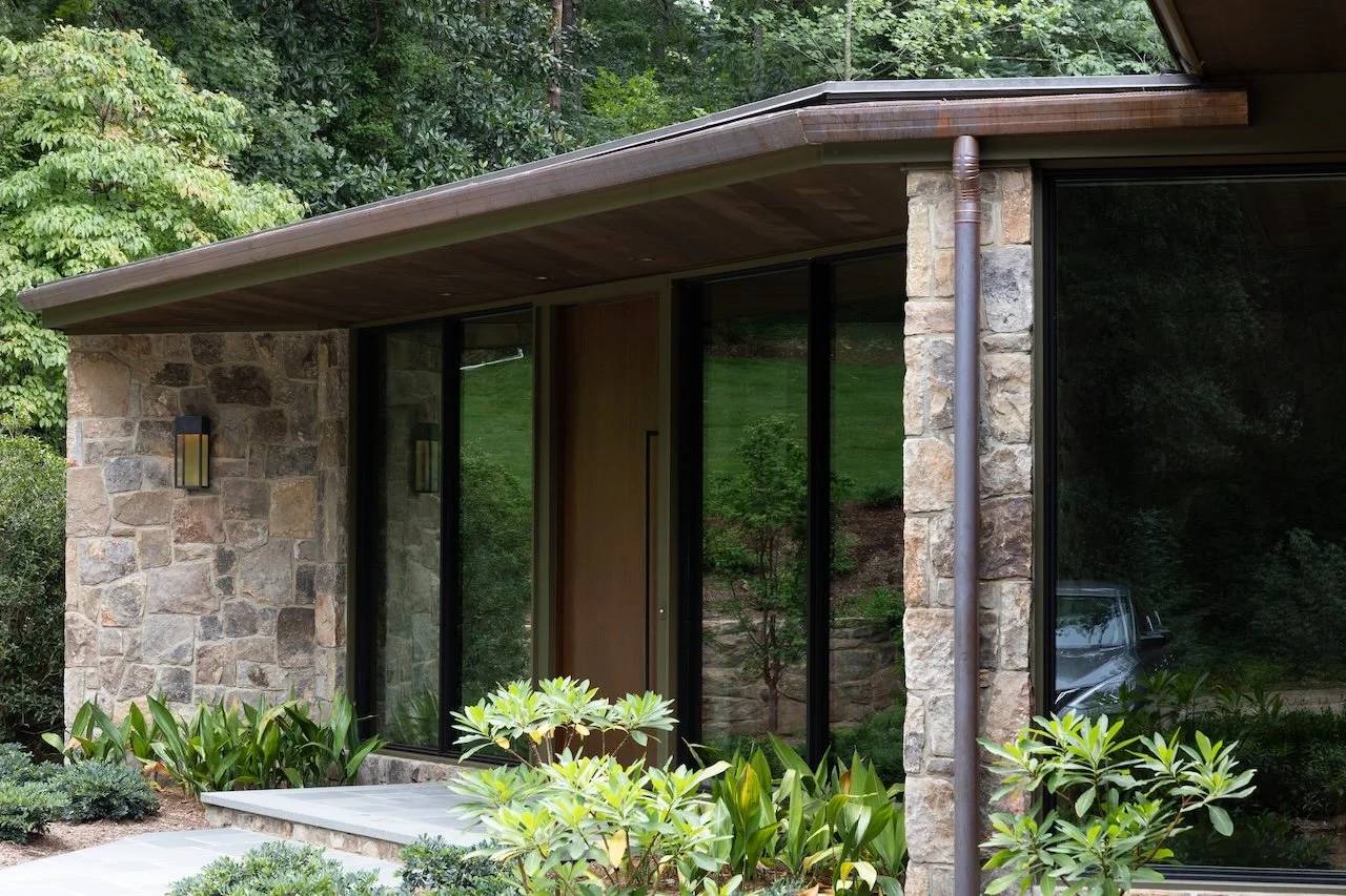 A modern house with stone and glass exterior, surrounded by green plants and trees.