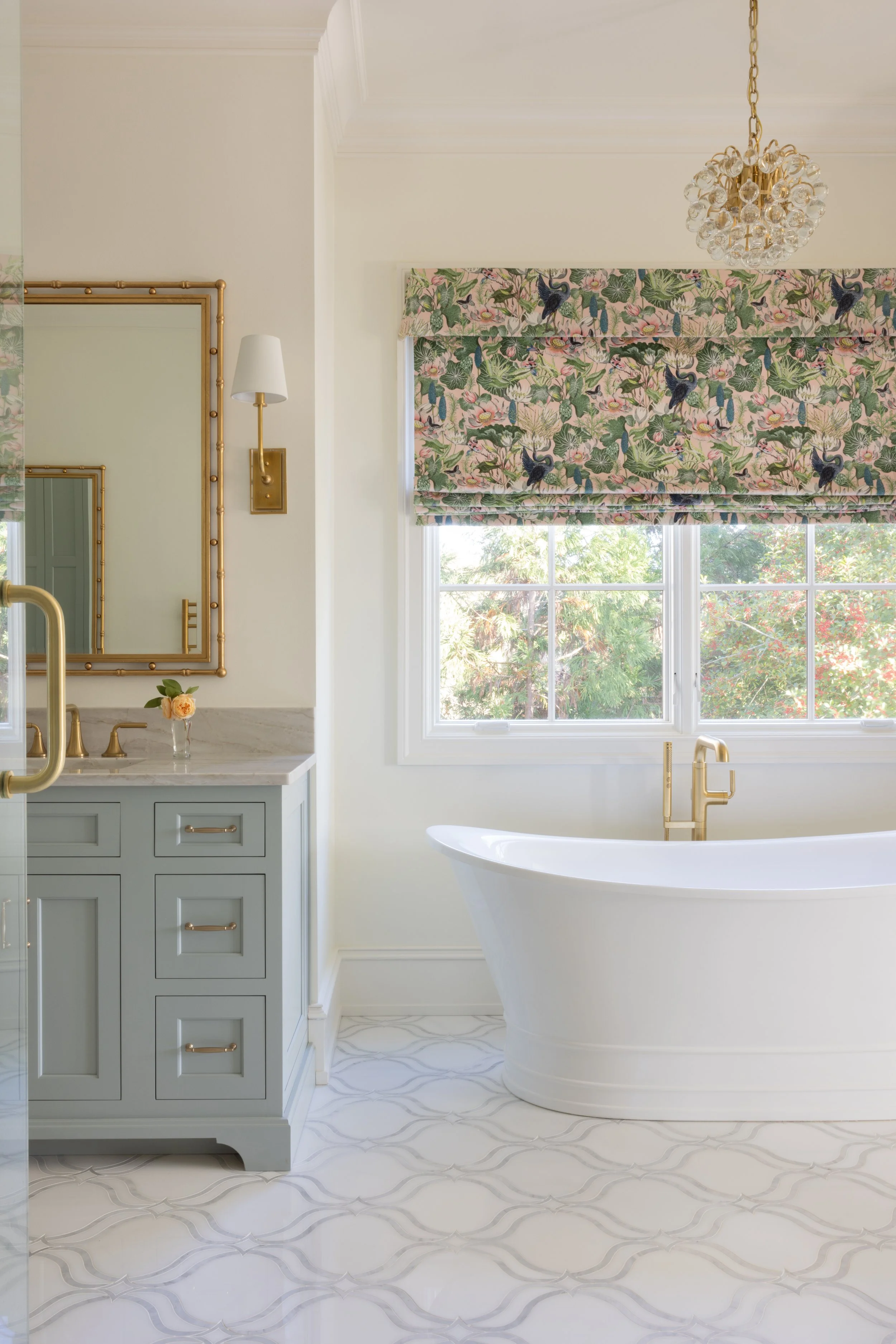 Bright bathroom featuring a white freestanding bathtub, a vanity with a marble countertop and light blue cabinetry, a mirror with a gold frame, a floral-patterned window shade, a gold chandelier, and a wall-mounted light fixture.