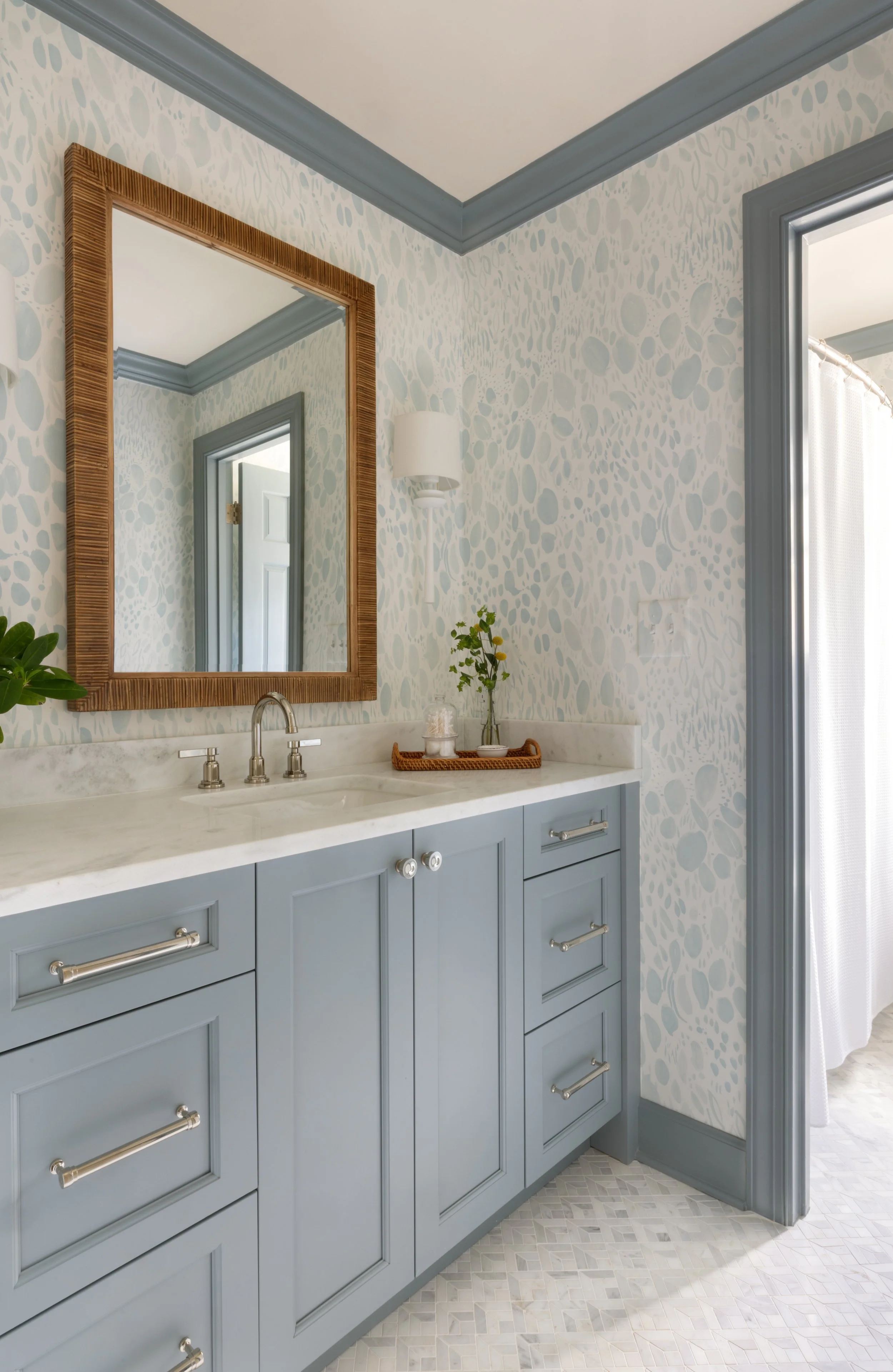 Bathroom vanity with blue cabinets, a white marble countertop, a wooden framed mirror, potted plant decor, and a doorway with a white curtain.