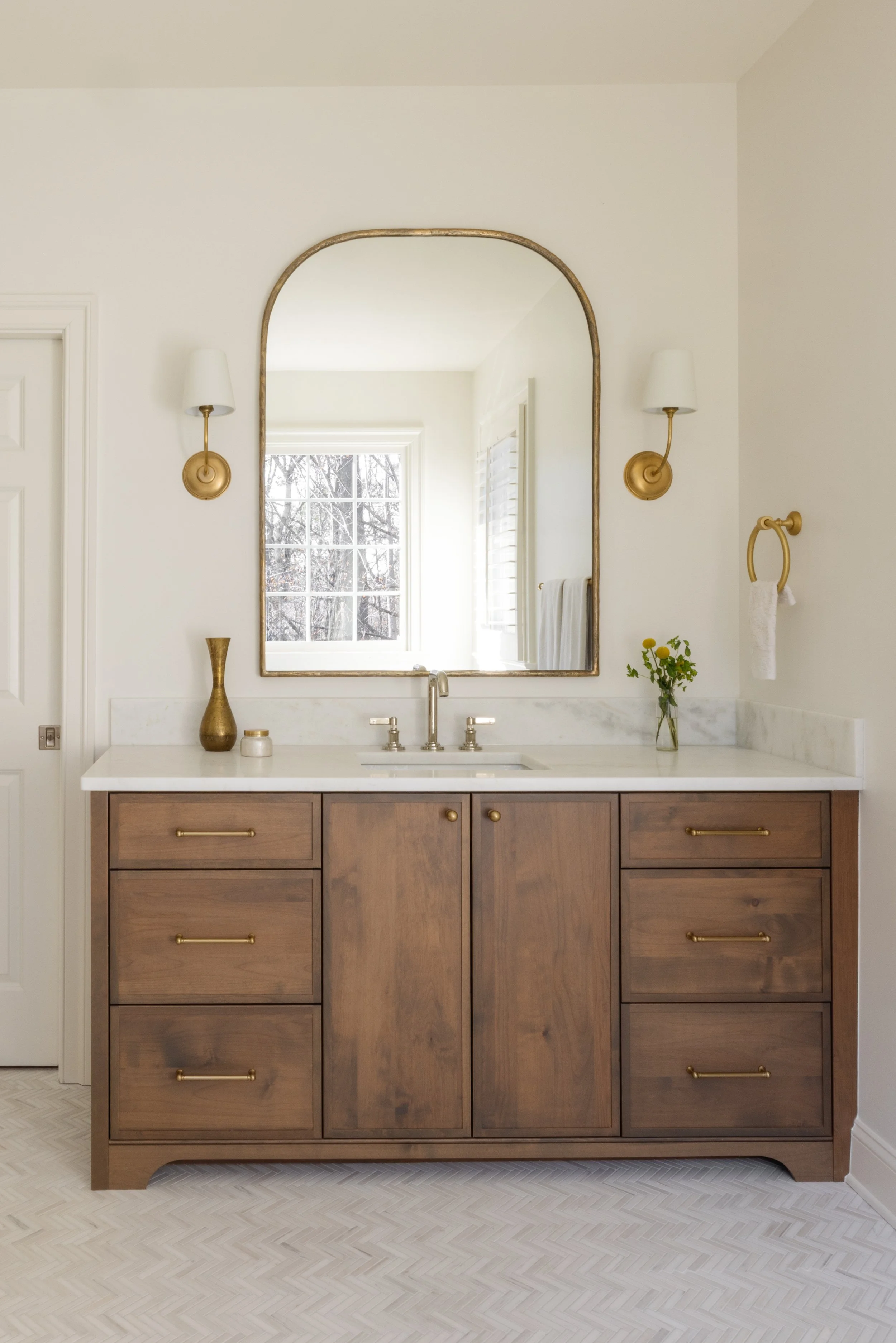 Bathroom vanity with a wooden cabinet, white marble top, white sink, gold fixtures, a large arched mirror, and decorative items including a gold vase and small container.