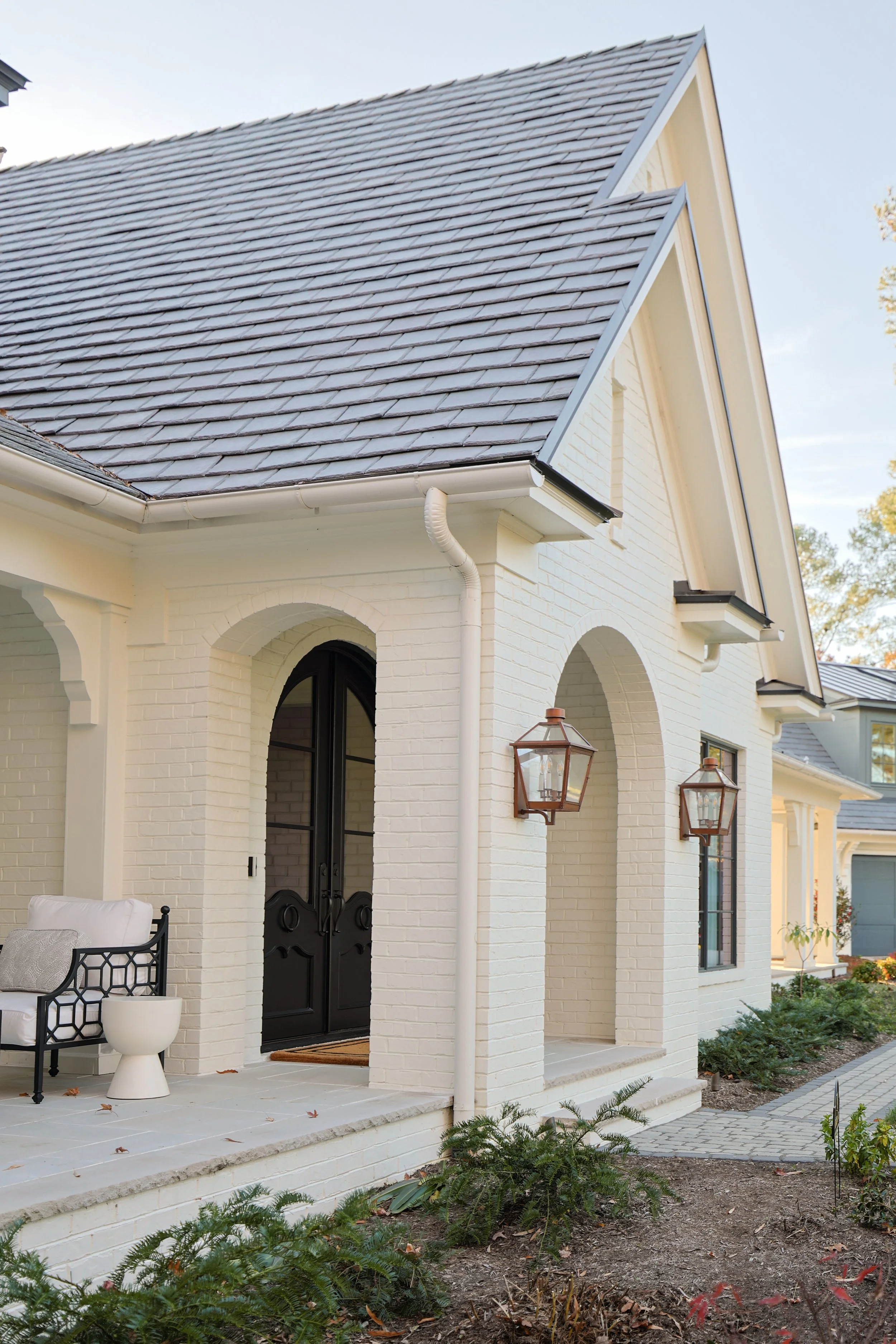 White brick house with black door, porch with outdoor furniture, arched windows, hanging lanterns, and garden.