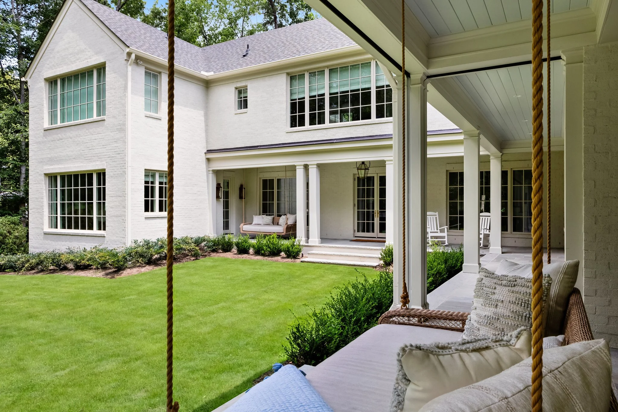View of a large white house with multiple windows, a porch with outdoor furniture, on a well-kept green lawn, seen from a covered patio with rocking chairs and pillows.