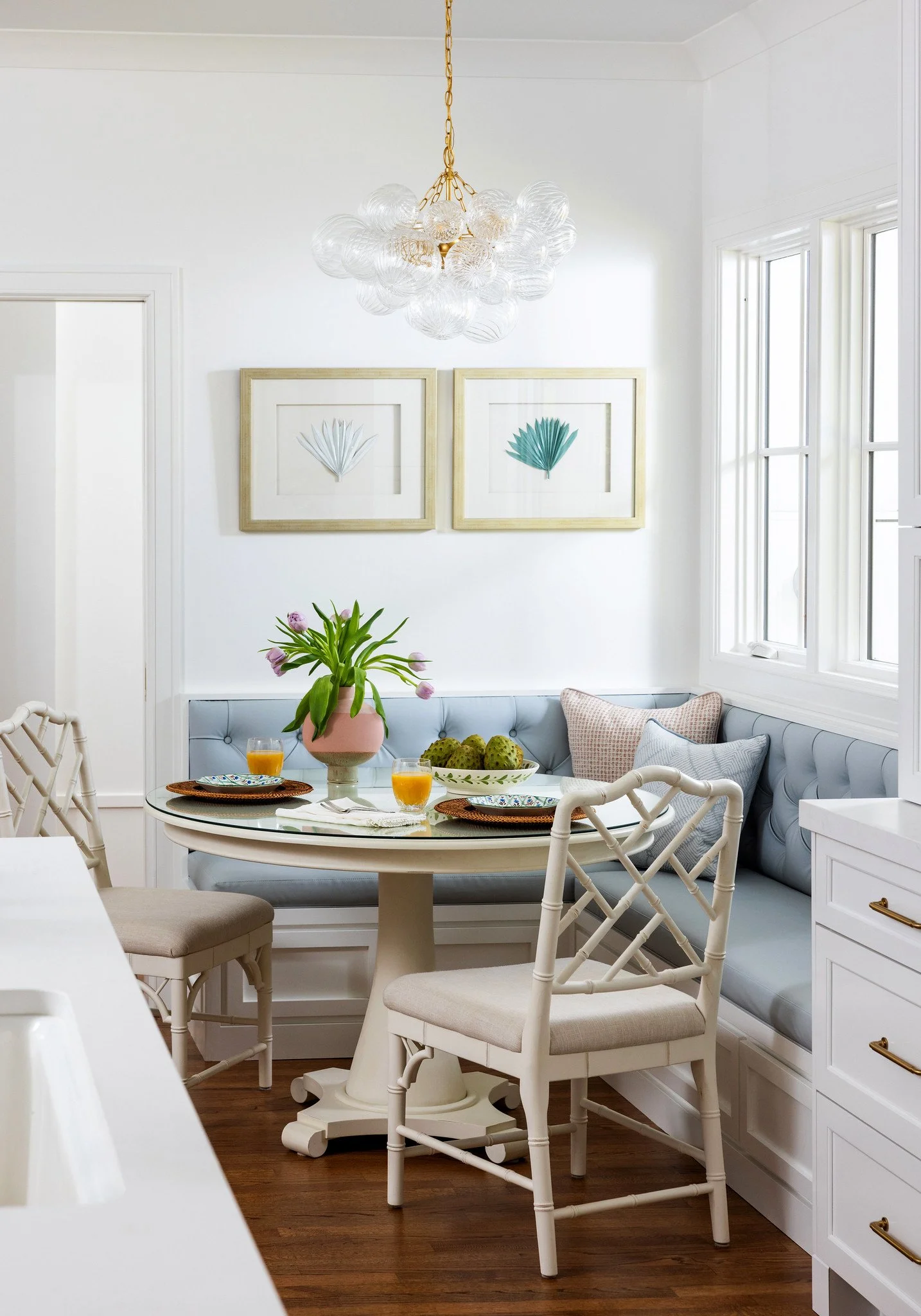 A cozy breakfast nook with a round table, a light blue cushioned bench, and white chairs. The table is set with placemats, plates, glasses of orange juice, and a pink vase with purple tulips. Decor includes framed botanical prints and a modern glass
