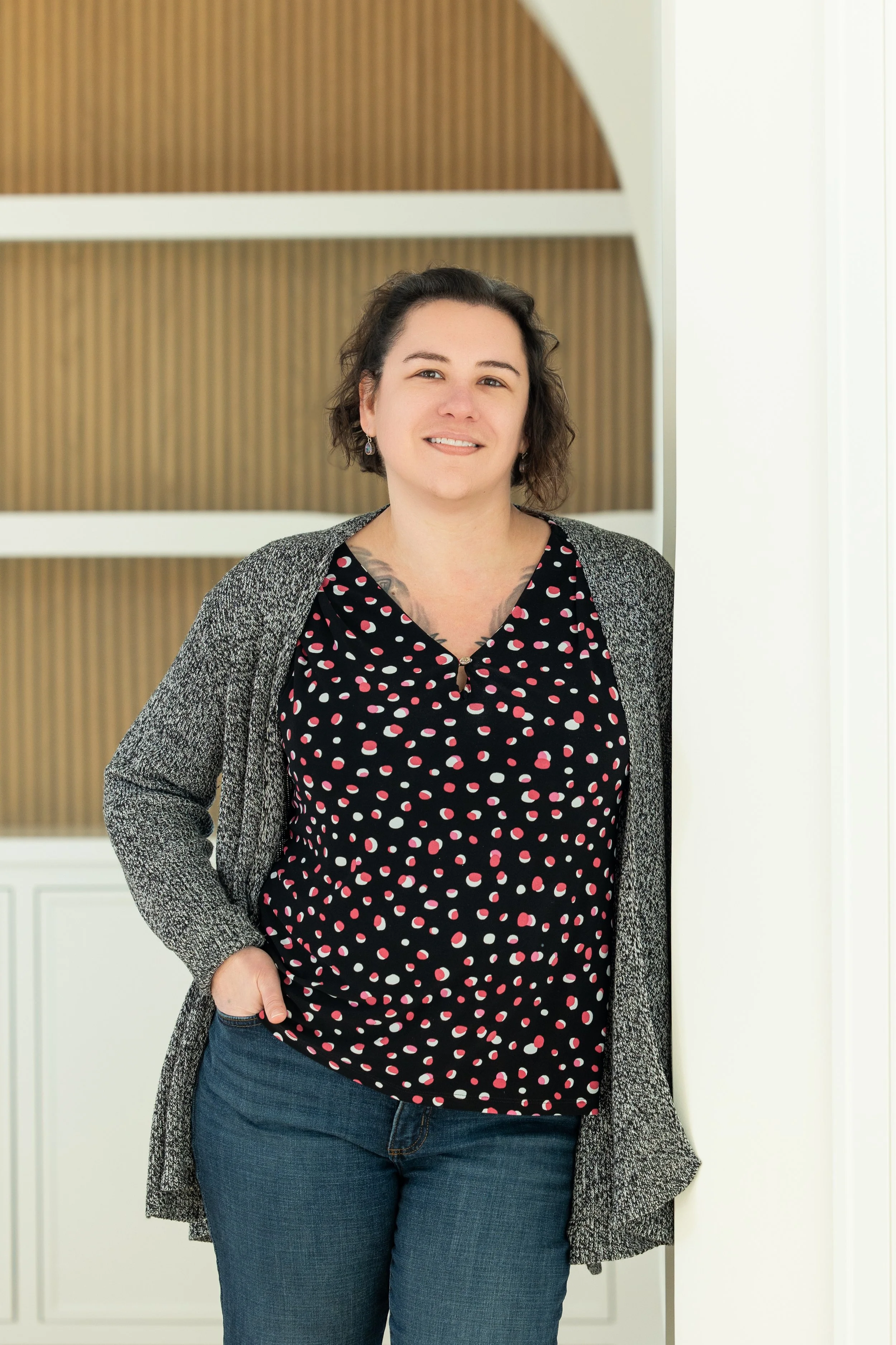 Smiling person with glasses and short curly hair wearing a blue top, standing against a wooden background.