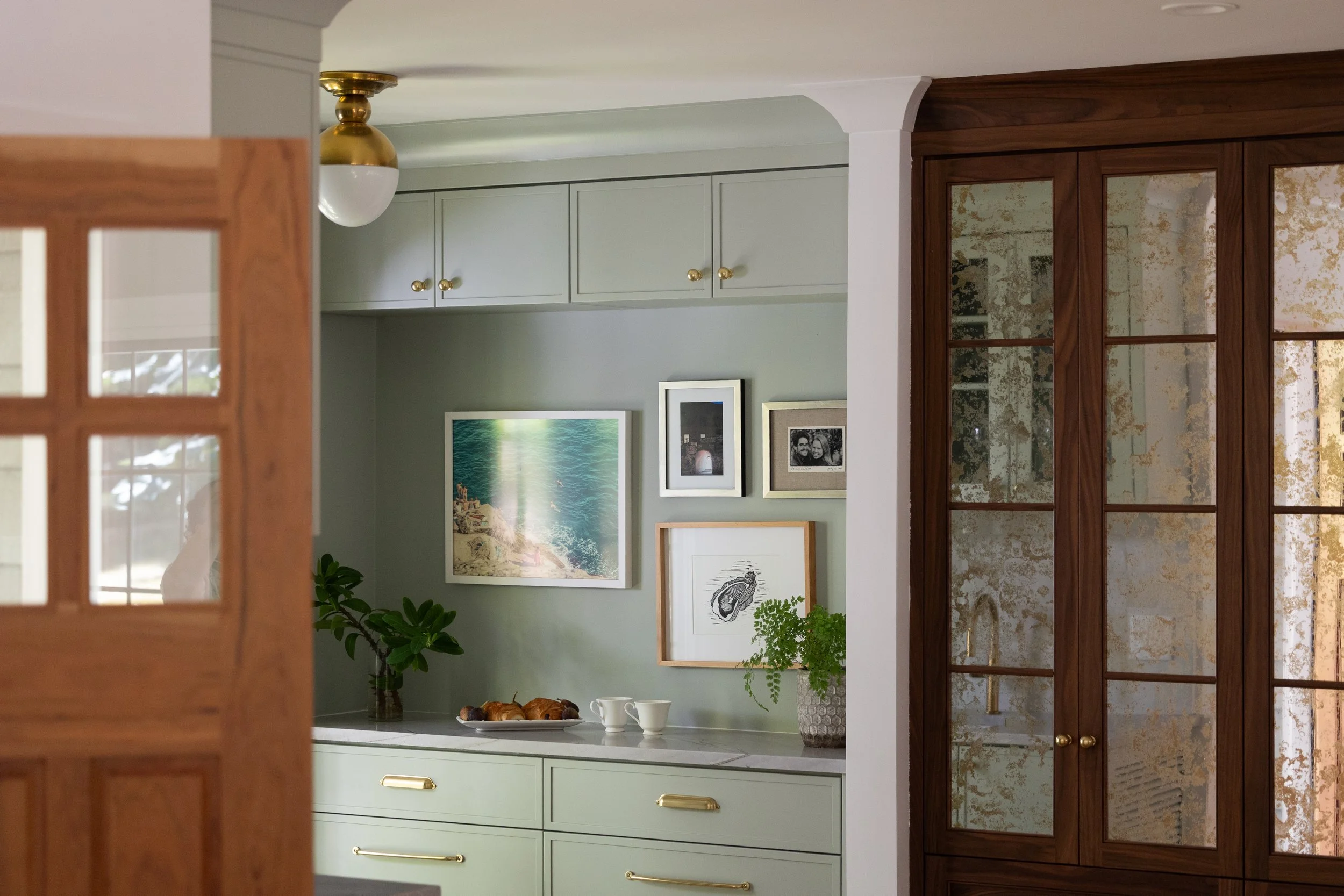 A cozy kitchen with light green cabinets, a white marble countertop, framed art on the wall, a potted plant, a plate of croissants, and two cups. A wooden door with glass panels partially blocks the view, and a wooden cabinet with glass doors is on t