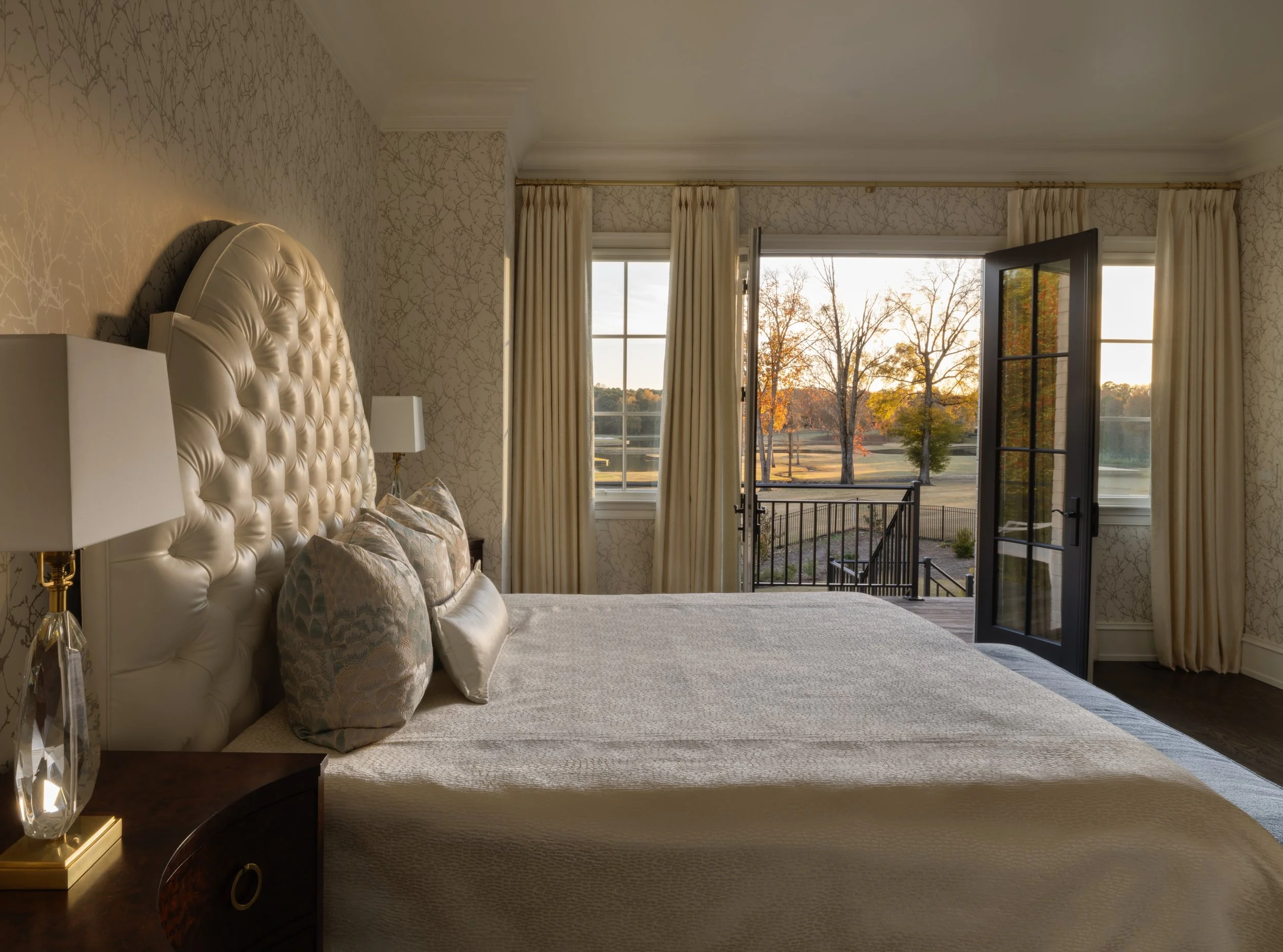A bedroom with an elegant, cream-colored tufted headboard, matching pillows, bedside lamps, cream curtains, and a glass door opening to a balcony with a view of trees and a park during sunset.