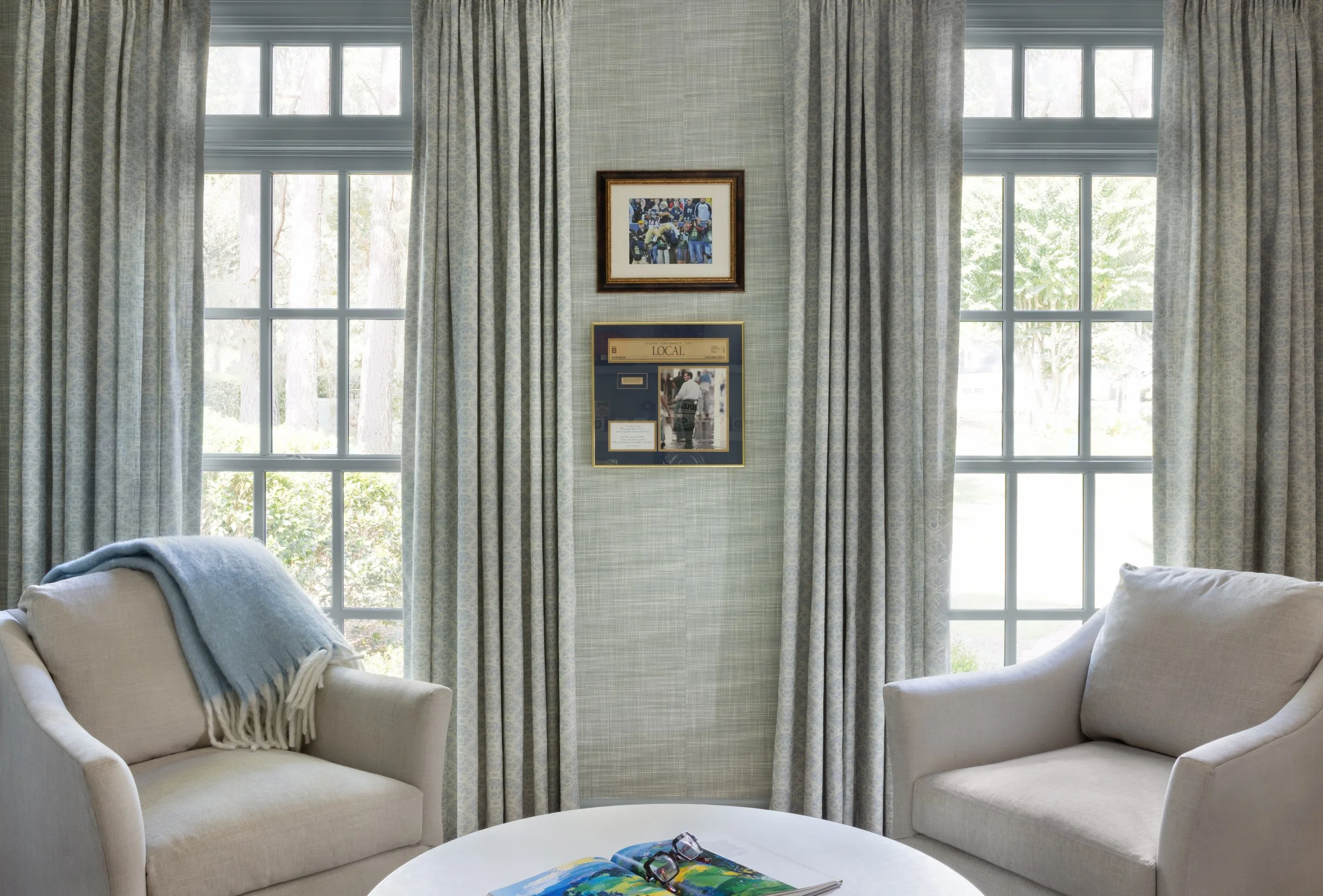 Living room with two beige armchairs, a white round coffee table with an open magazine and glasses, large windows with light patterned curtains, and framed pictures on a wall between the windows.