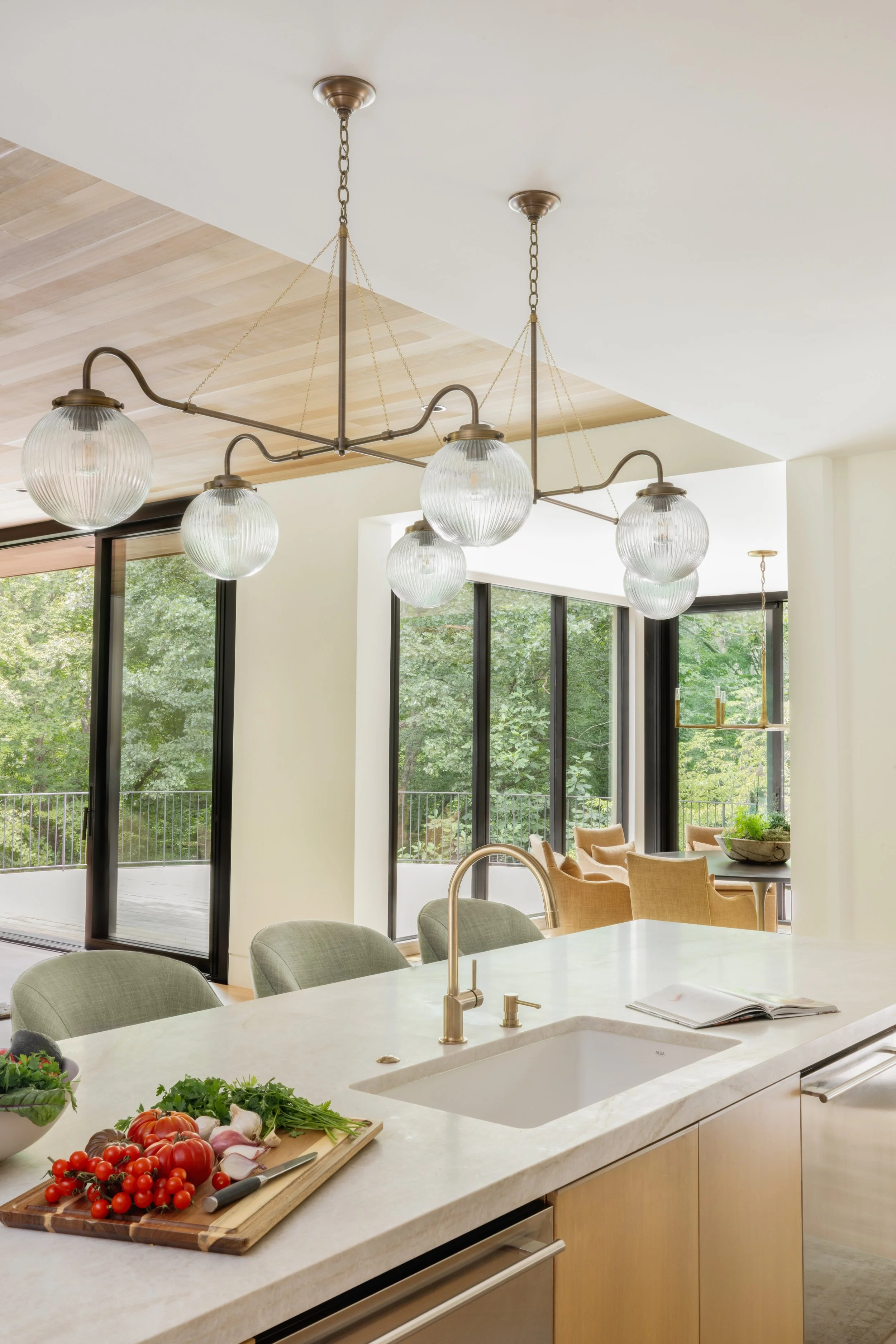 Modern kitchen with white marble island, fresh vegetables, and a large glass window overlooking greenery.