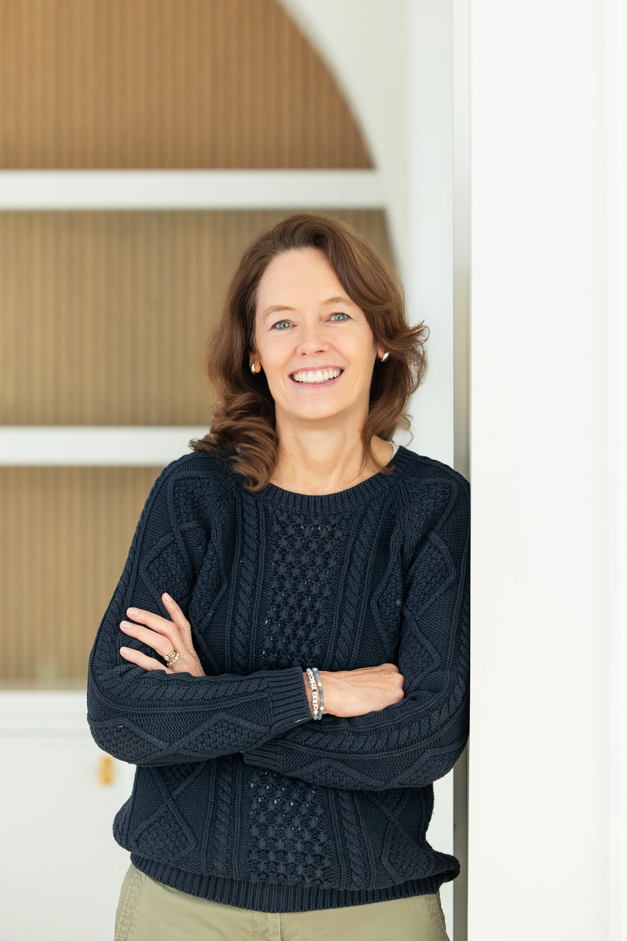 A woman with curly hair is smiling while holding a phone to her ear. She's wearing a light blue blouse. In the background, there are sketches and a textured wall.