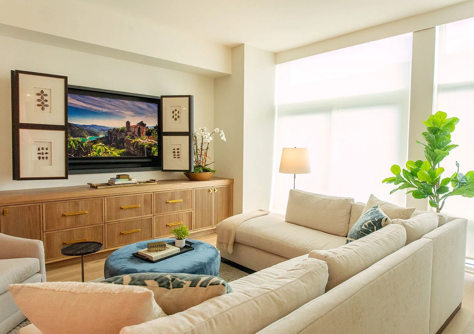 Bright living room with white sofa, large window with sheer curtains, wooden sideboard, framed landscape art, television, potted plant, floor lamp, and decorative pillows.