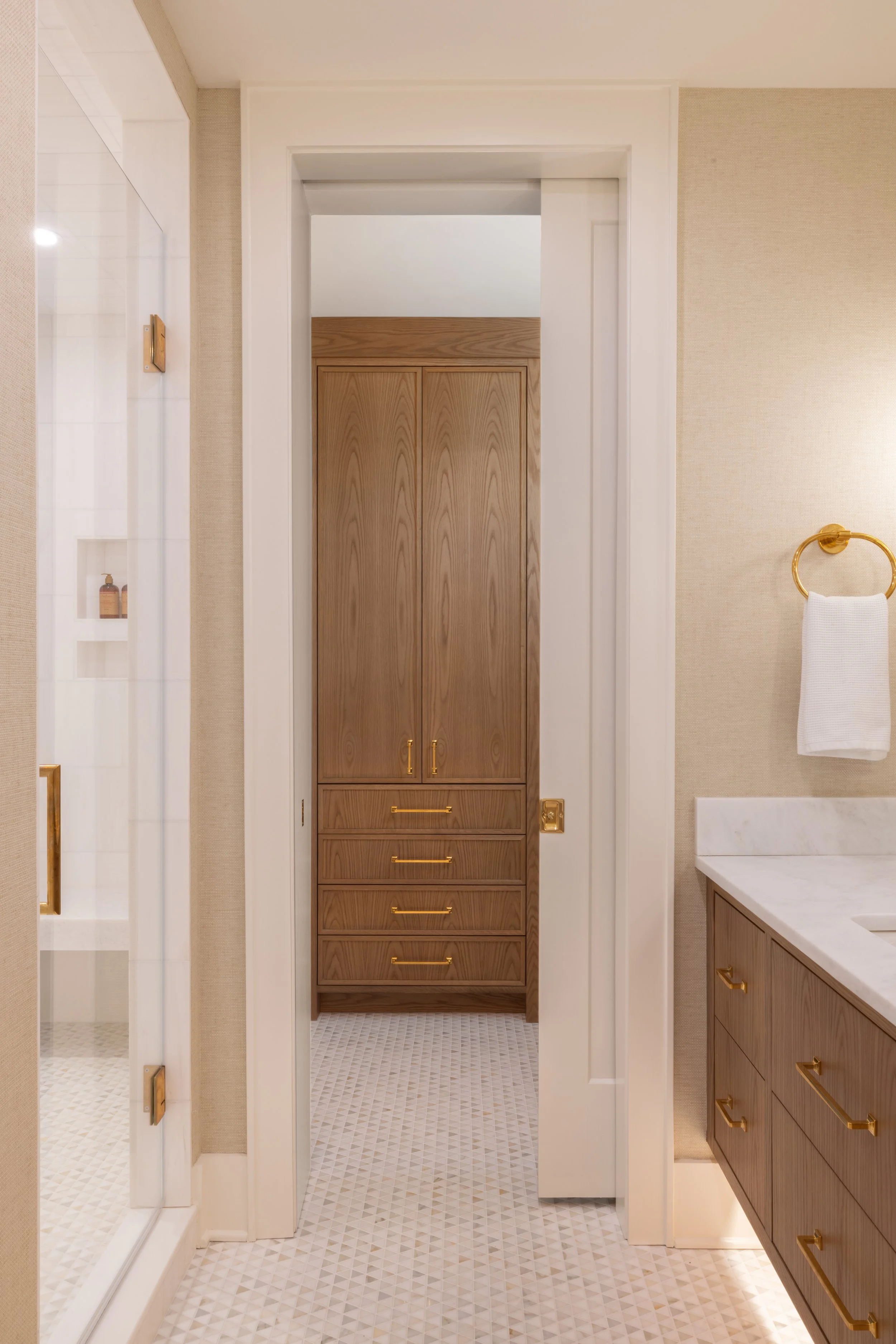 View of a bathroom with a wooden cabinet with gold handles at the end of a small hallway, beige walls, a white countertop on the right, a towel ring with a white towel, and part of a glass shower on the left.