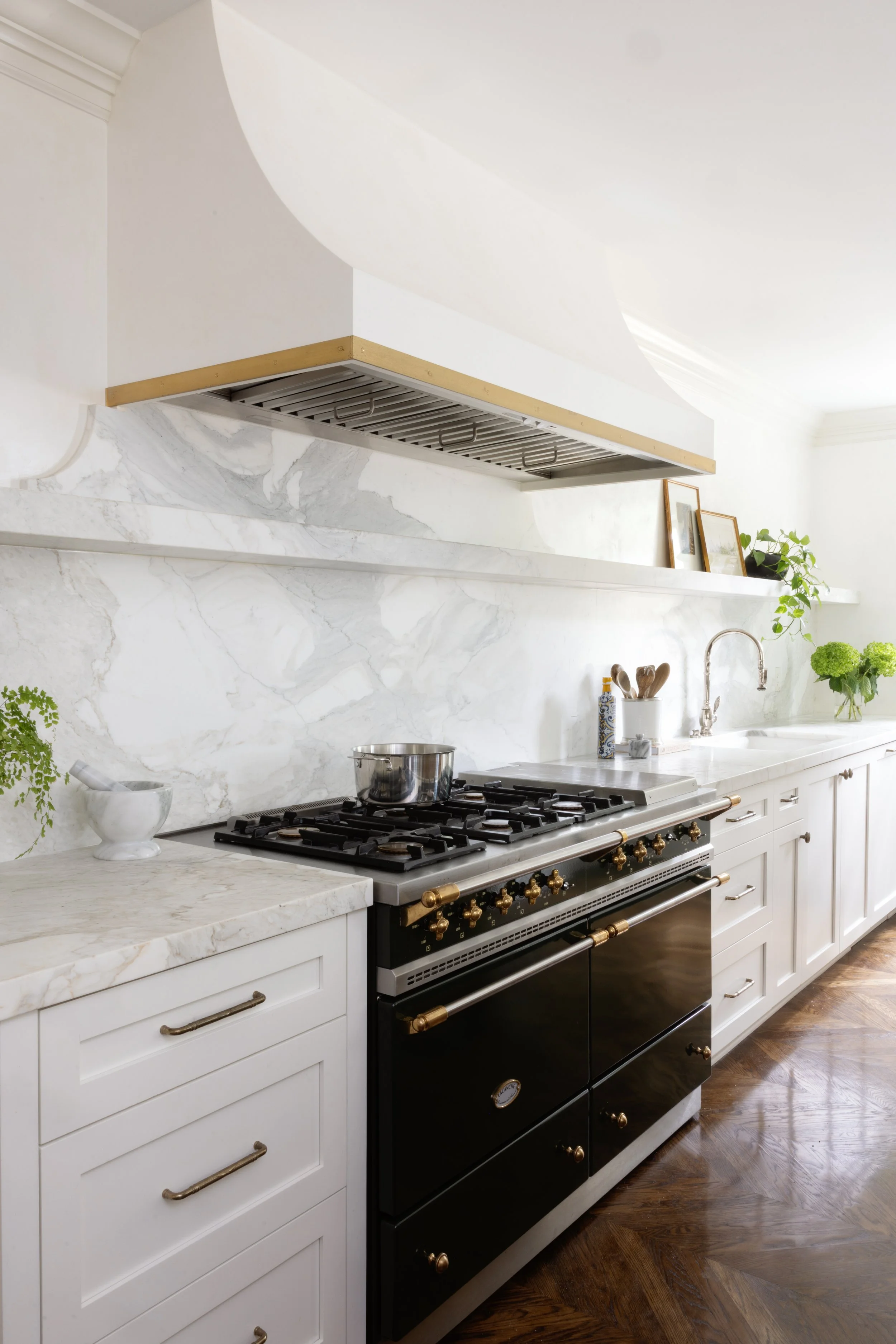 A kitchen with white cabinetry, a black and gold stove, marble countertops and backsplash, and a gold range hood. There are green plants and framed pictures on the shelves.