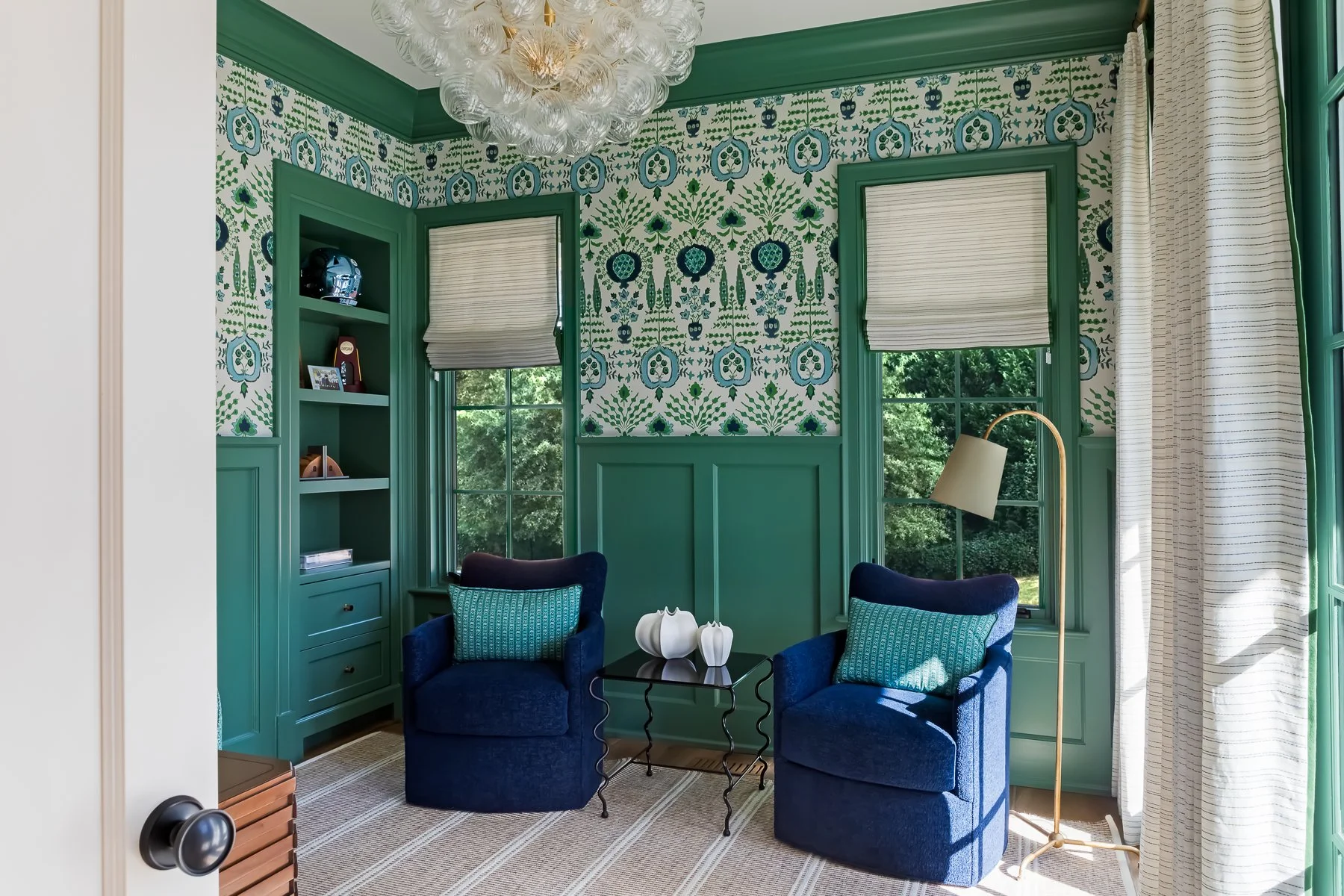 Cozy reading nook with blue chairs, patterned wallpaper, green wainscoting, decorative shelves, a floor lamp, and a small glass table with white vases.